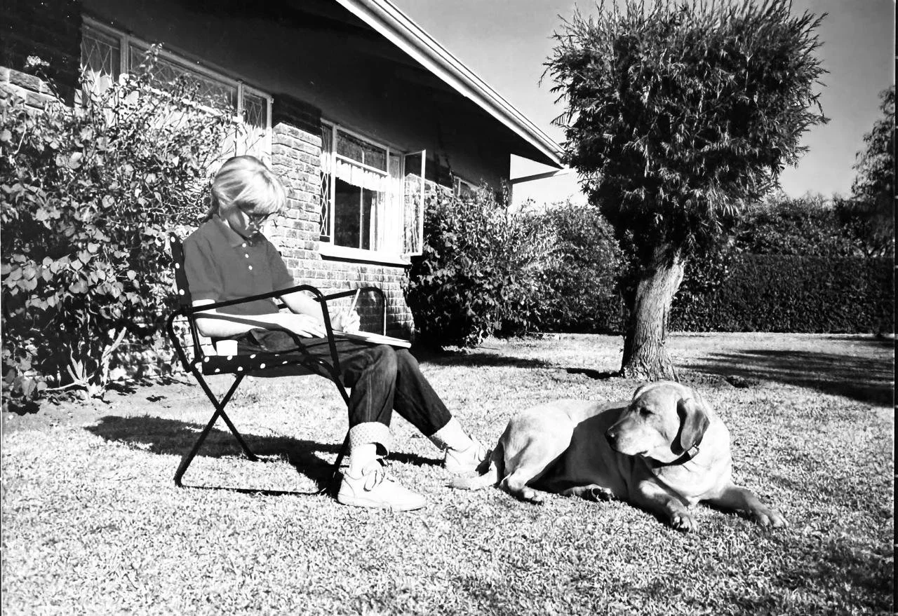 A girl sitting on a garden chair reading a book next to a lying dog in a yard with trees and bushes, black-and-white photo.