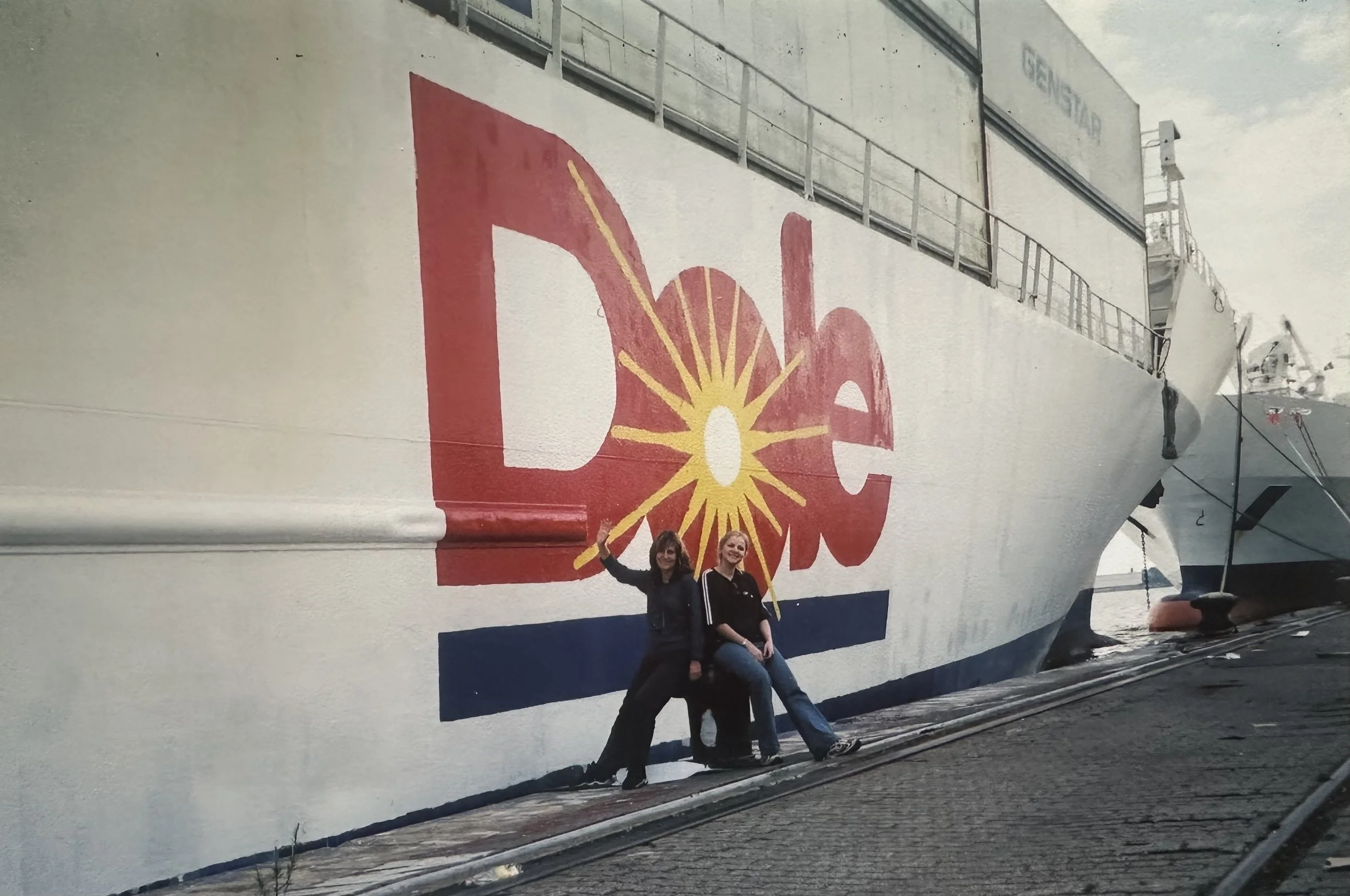 Two women standing in front of a large ship with painted red, yellow, and white design and the word "Dole" on it.