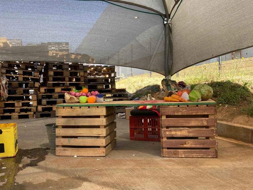 Market stand made of wooden pallets with produce including apples, carrots, lettuce, and cabbage, under a shadecloth in an outdoor setting.