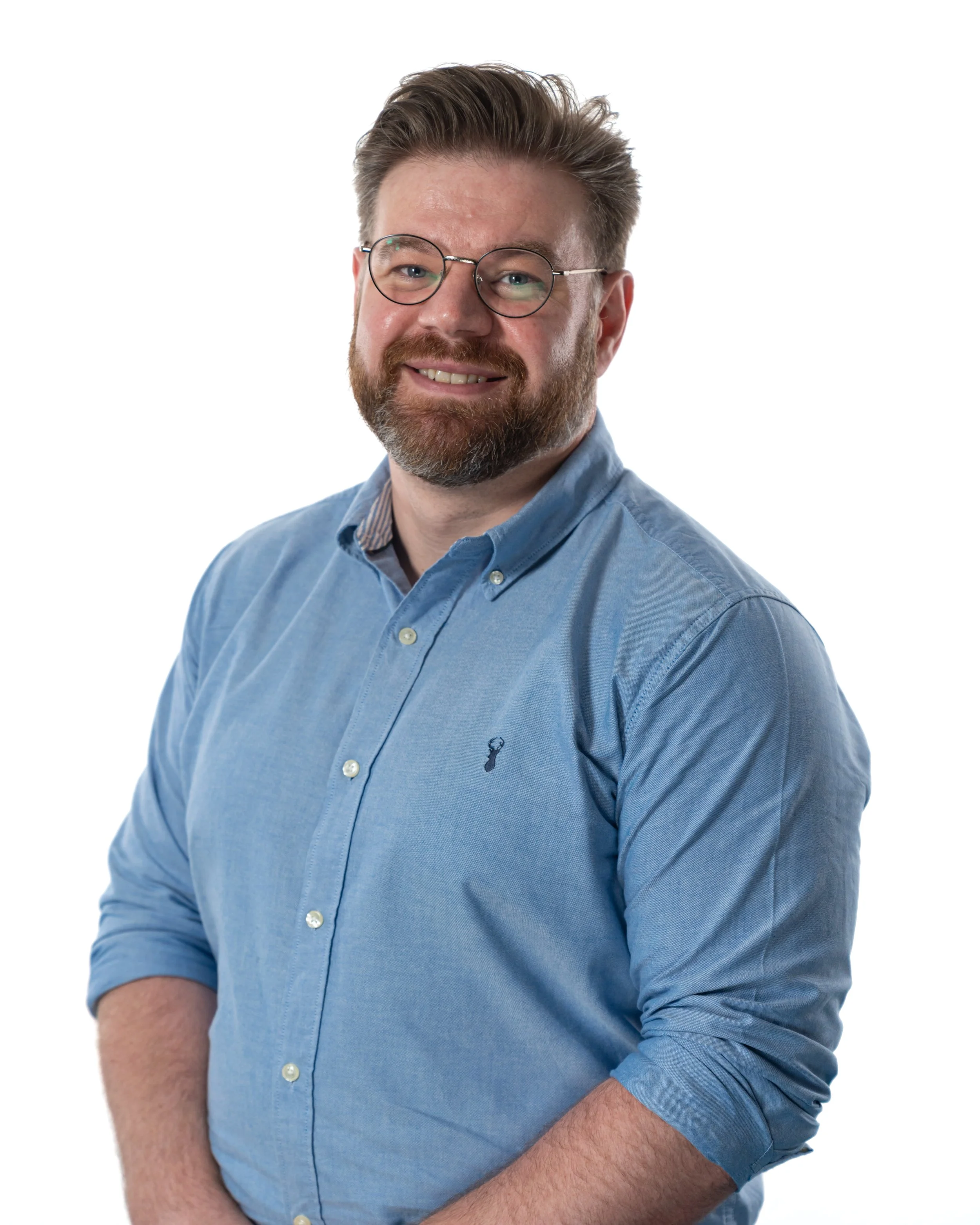 A man with brown hair, glasses, and a beard wearing a blue button-up shirt, smiling, against a white background.