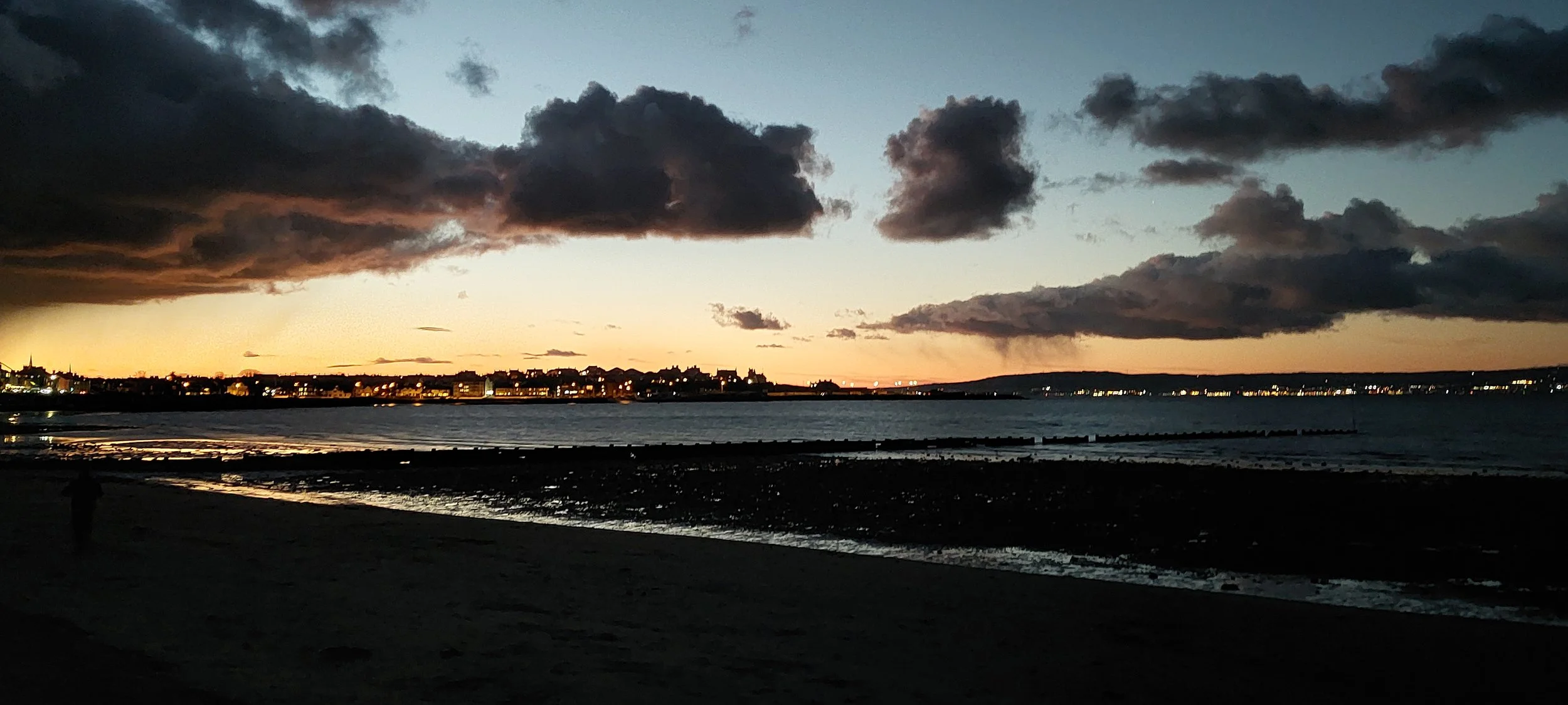 Sunset over a beach with dark clouds in the sky and city lights along the shoreline.