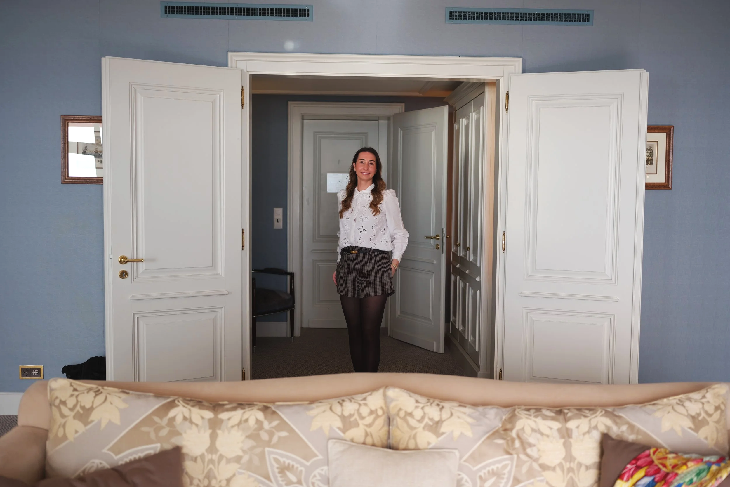 Une femme souriante se tient dans une pièce, devant une porte ouverte, avec un mur bleu derrière elle. Elle porte une chemise blanche, une jupe à carreaux et des collants noirs, et a les cheveux longs et ondulés.