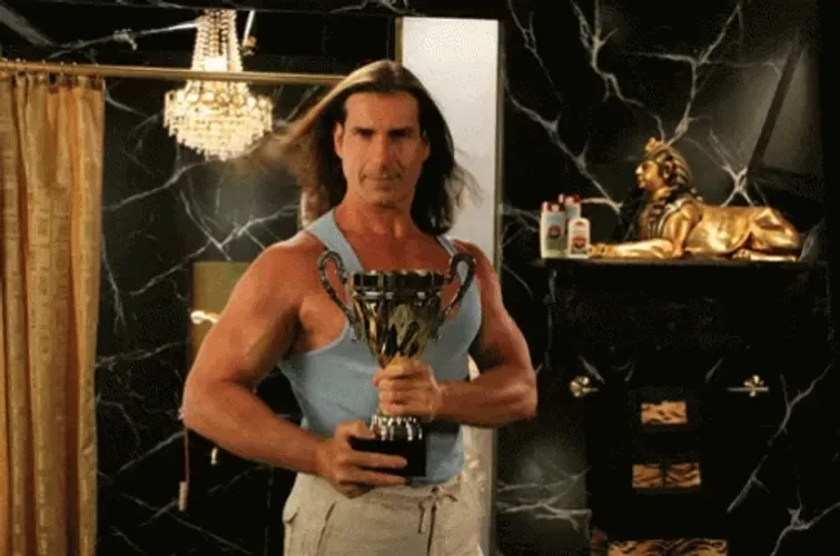 Man with long hair wearing a sleeveless blue shirt, holding a trophy, standing in a room with black marble walls, a chandelier, and decorative items including a golden lion statue.