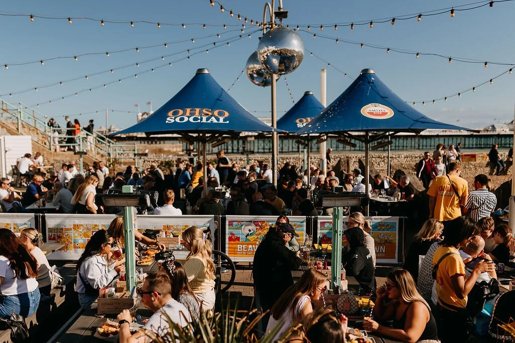 A crowded outdoor event on a sunny day with blue tents labeled "OHIO SOCIAL," string lights overhead, and people seated at tables and standing, enjoying food and drinks.