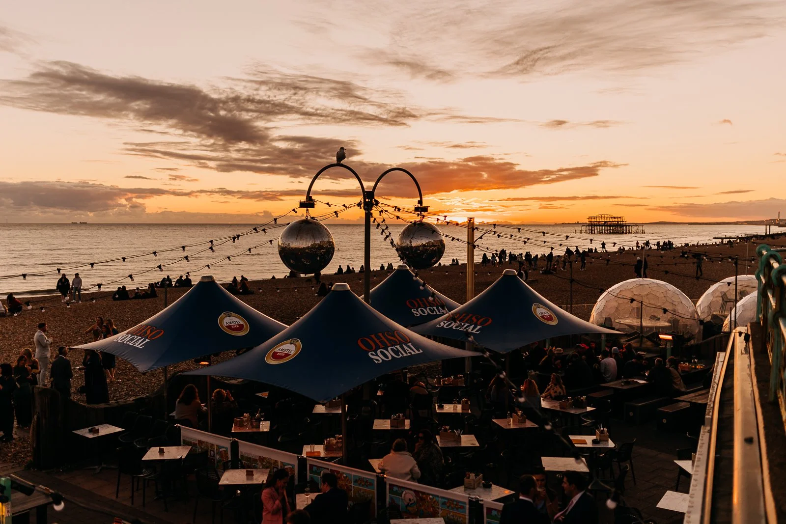 People gathered at an outdoor beachside restaurant with blue umbrellas that read 'OHSO SOCIAL' during sunset. The sky is orange with scattered clouds, and the calm ocean and a pier are visible in the background.
