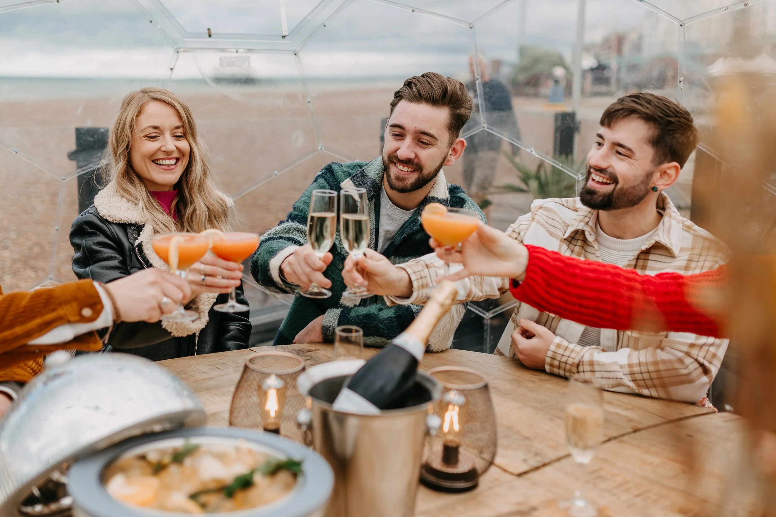 Group of friends celebrating with drinks inside a greenhouse or glass enclosure, surrounded by food and decor.