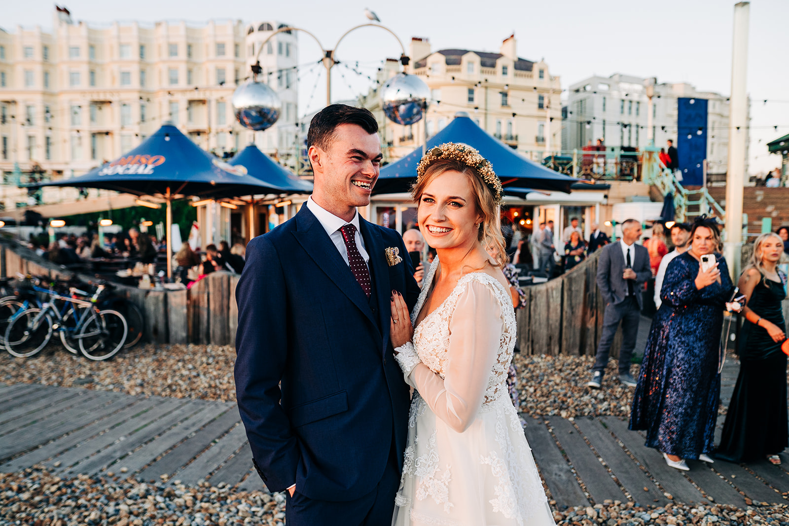 A bride and groom smiling together at an outdoor wedding reception during sunset, with guests and seaside buildings in the background.