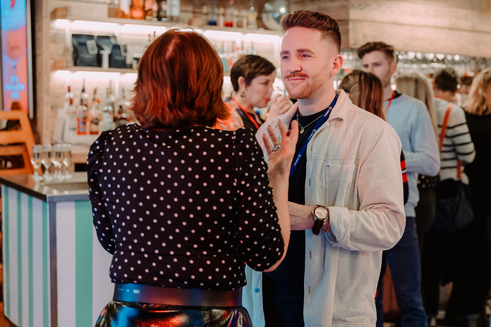 A man and woman engaged in a conversation at a bar or social event, with other people in the background.