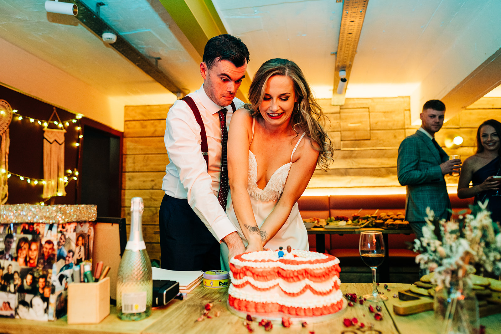 A man and woman are cutting a birthday cake at a celebration, surrounded by friends and decorations.