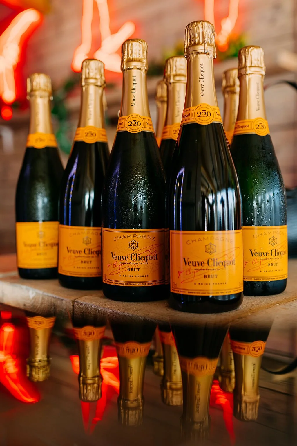 Six bottles of Veuve Clicquot champagne on a wooden surface with a mirror reflection, a brick wall, and neon decorations in the background.