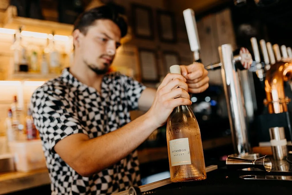 A man in a patterned shirt opening a bottle of wine at a bar or brewery.