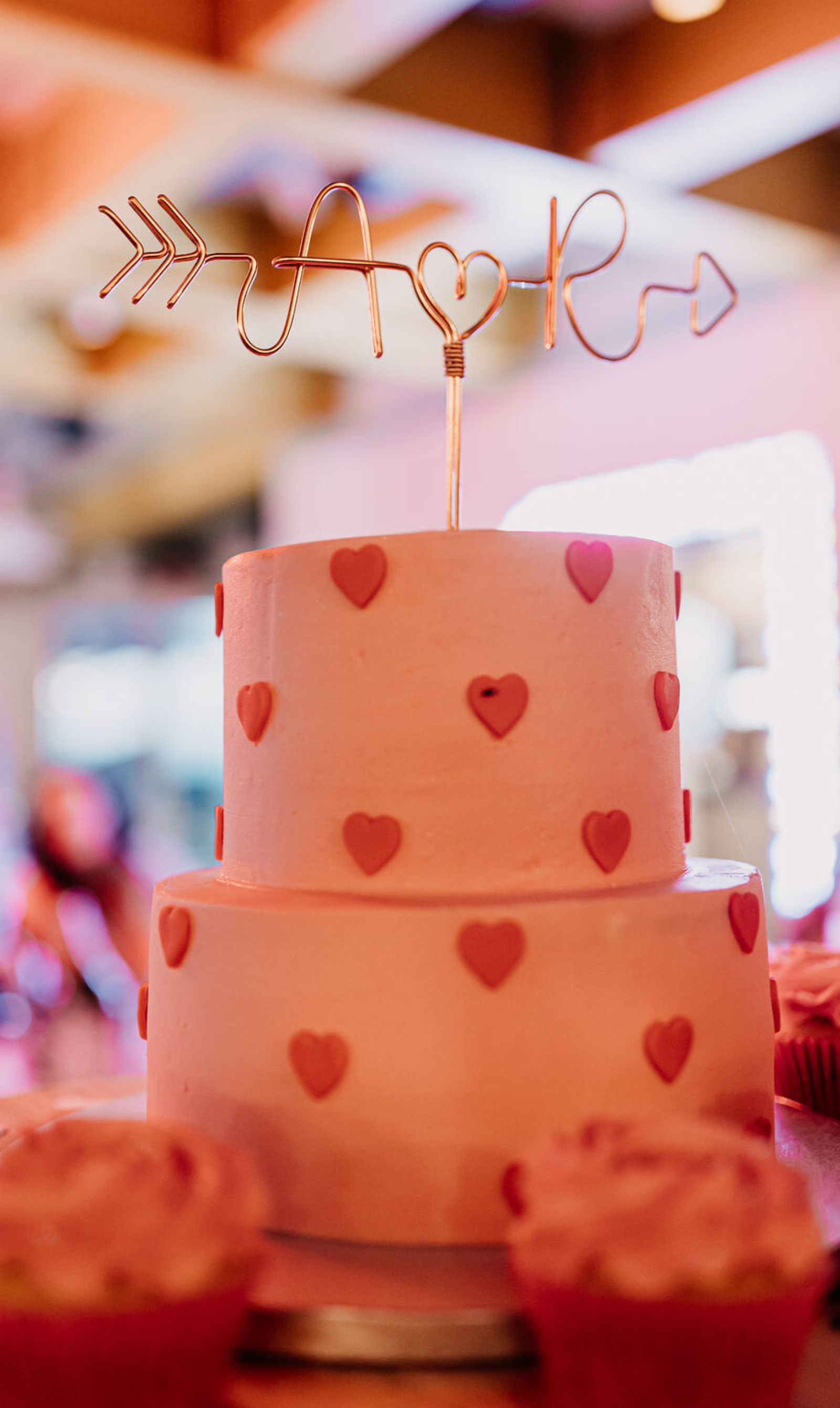 A pink, multi-tiered cake decorated with small red hearts, with a wire cake topper spelling 'A♡R' with arrows and hearts, against a blurred background of pink and warm tones.