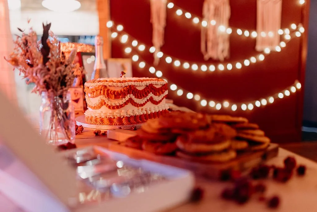 A birthday or celebration table with a decorated cake, plate of cookies, and dried flowers, with warm string lights and hanging decorations in the background.