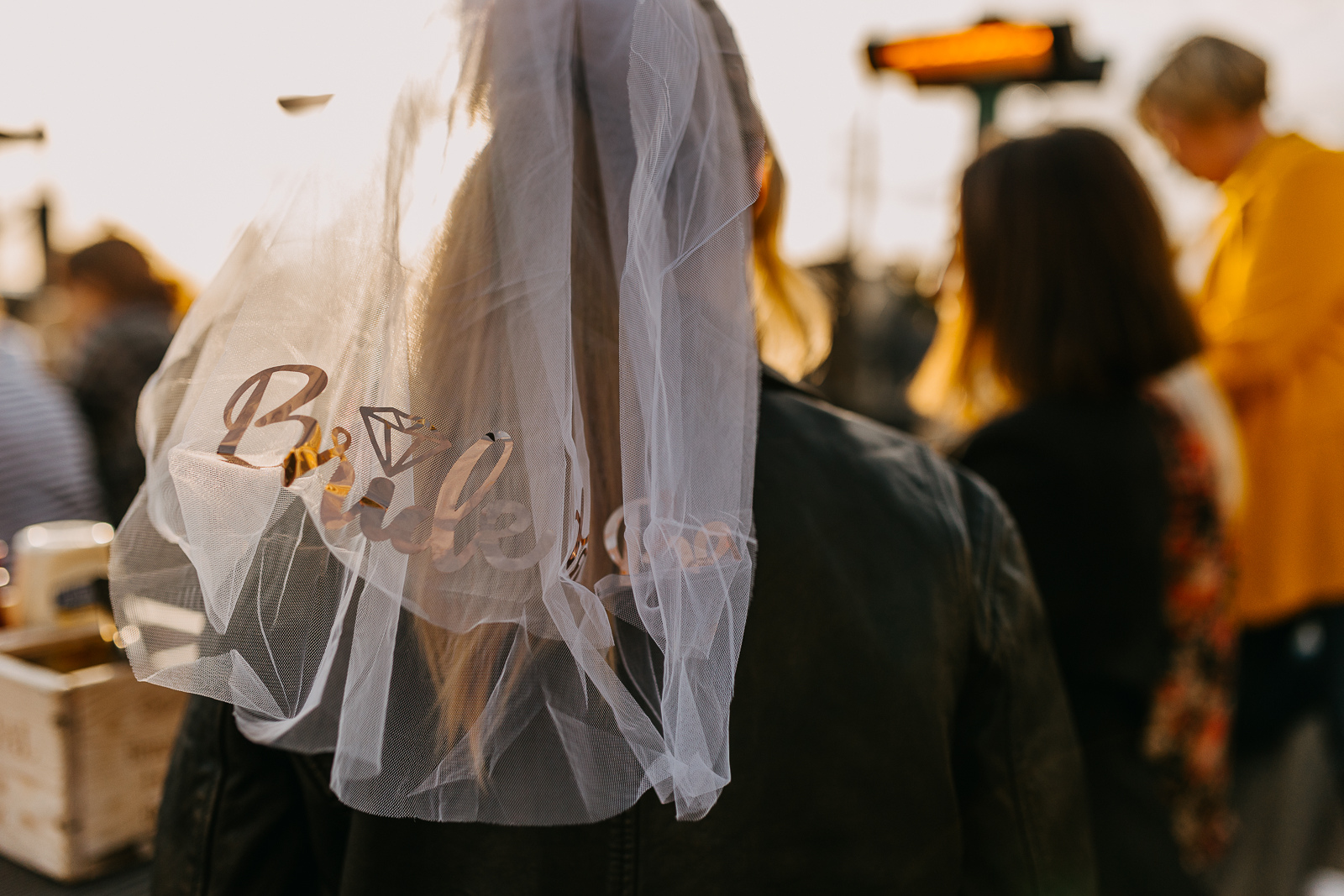 A person wearing a veil with decorative items attached, standing among a crowd of people at an outdoor event during sunset.