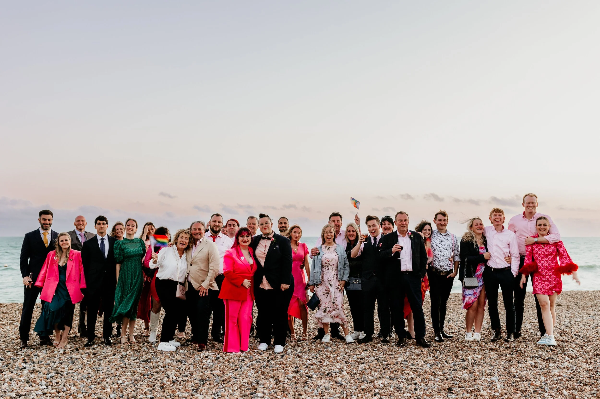 A group of smiling people dressed in colorful attire, standing on a rocky beach with the ocean and sky in the background, enjoying a celebration at sunset.