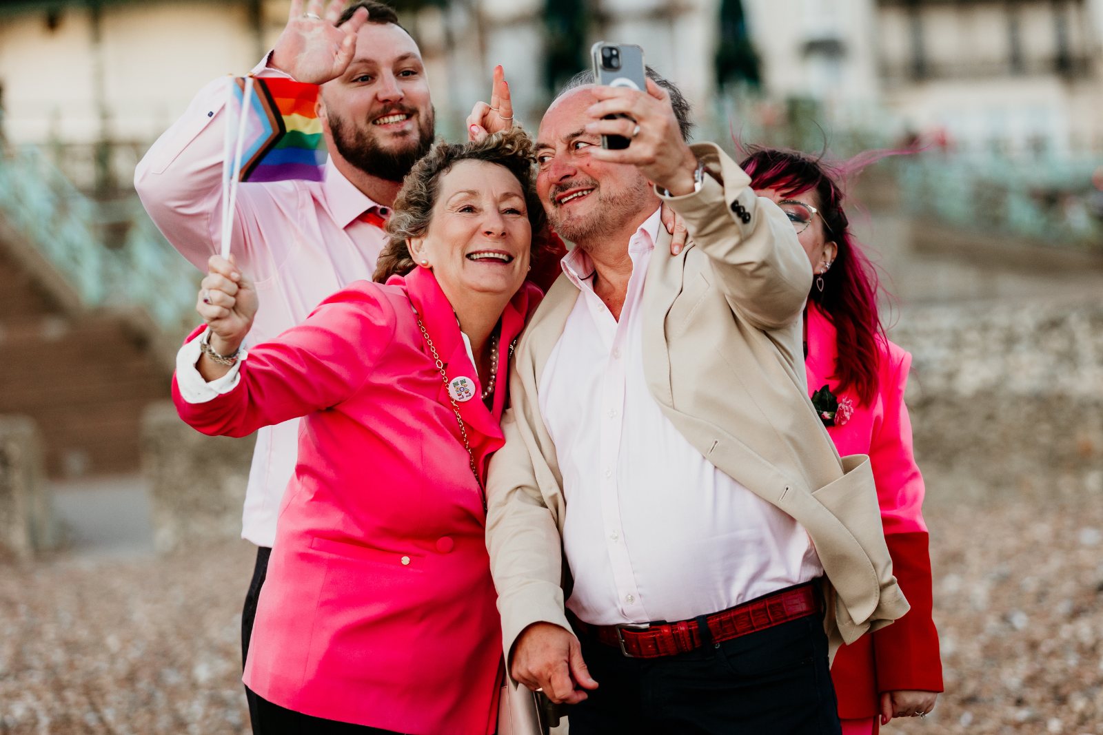 A group of five diverse adults taking a selfie outdoors. They are smiling and appear to be celebrating, with some wearing pink and red clothing and a rainbow pride flag visible.