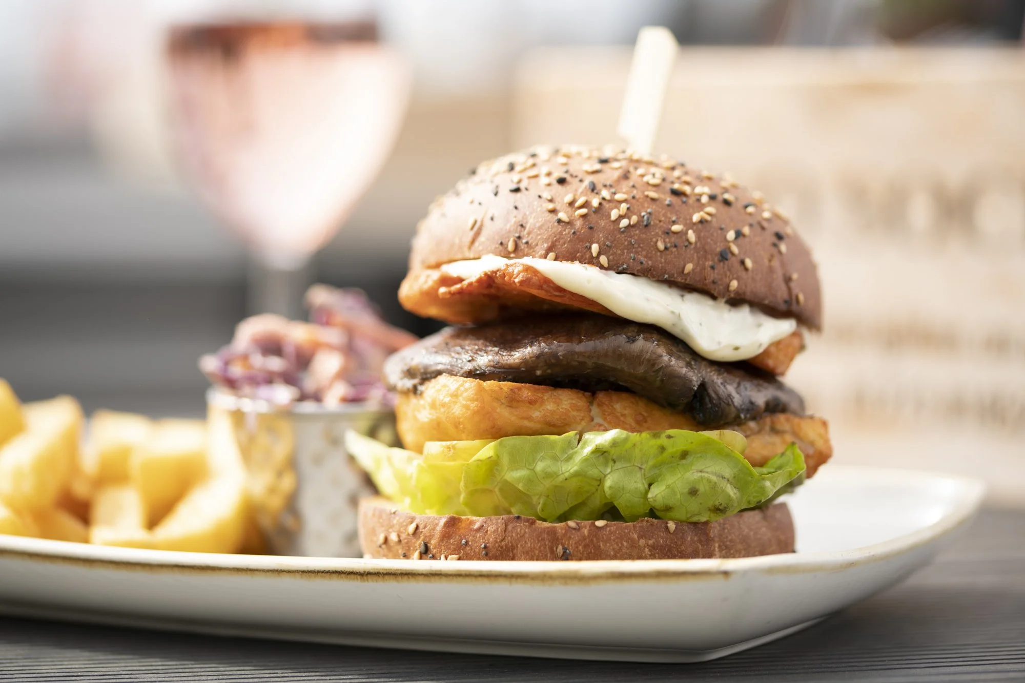 A close-up of a multi-layer burger with lettuce, tomato, cheese, grilled chicken, a fried chicken patty, a beef patty, bacon, mayonnaise, and a sesame seed bun, served on a white plate with a side of fries and a beverage in a blurred background.