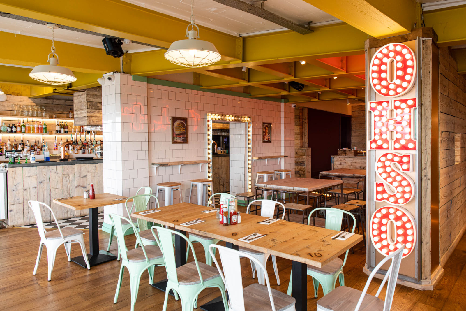 Interior of a trendy restaurant or bar with wooden tables and metal chairs, a white-tiled bar area, decorative lighting, and a large illuminated sign that says "OPEN".