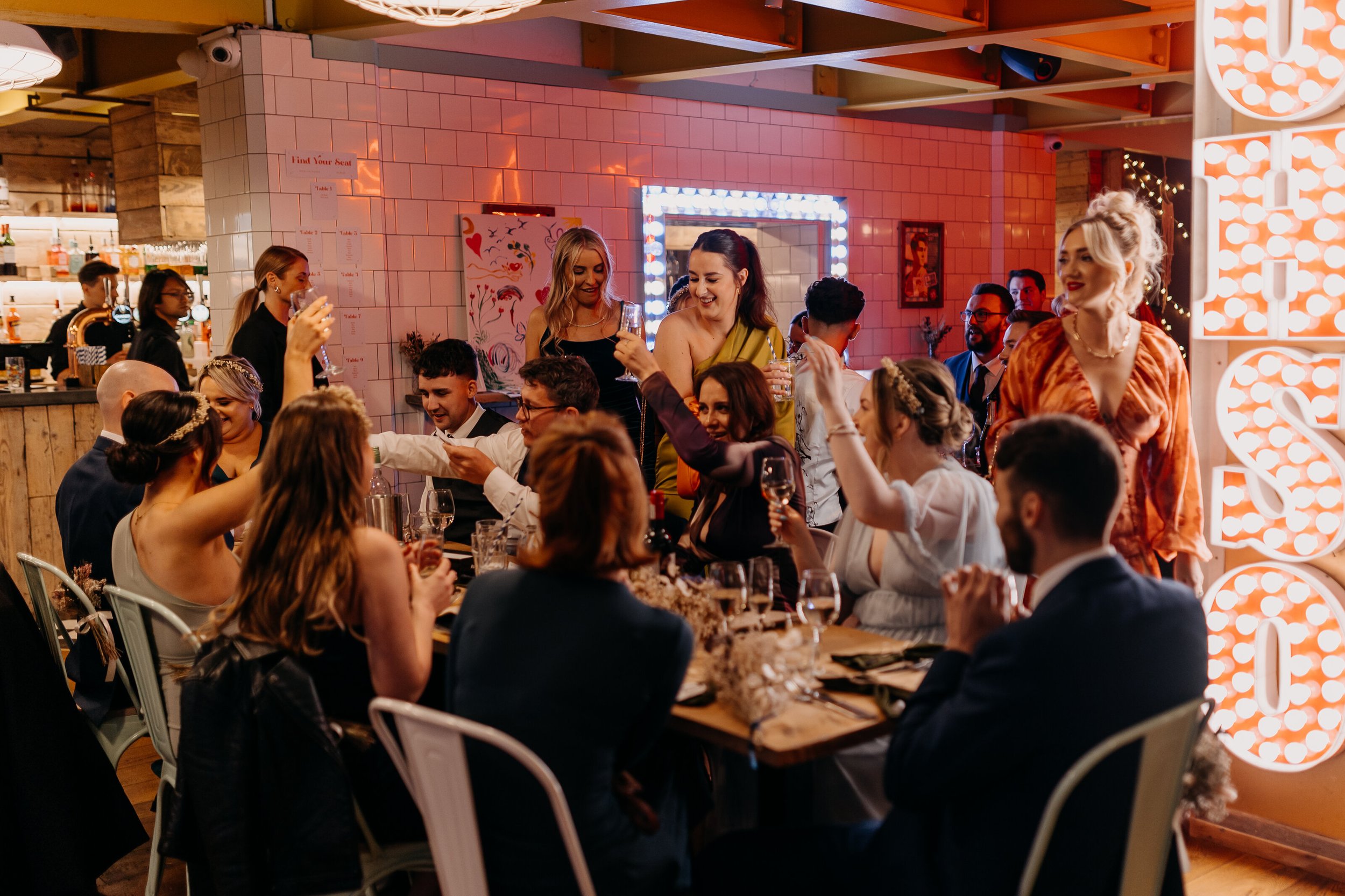 A lively indoor celebration with people sitting at a long table, some standing and smiling, in a warmly lit setting with decorative lighting, artwork on the walls, and a bar area in the background.