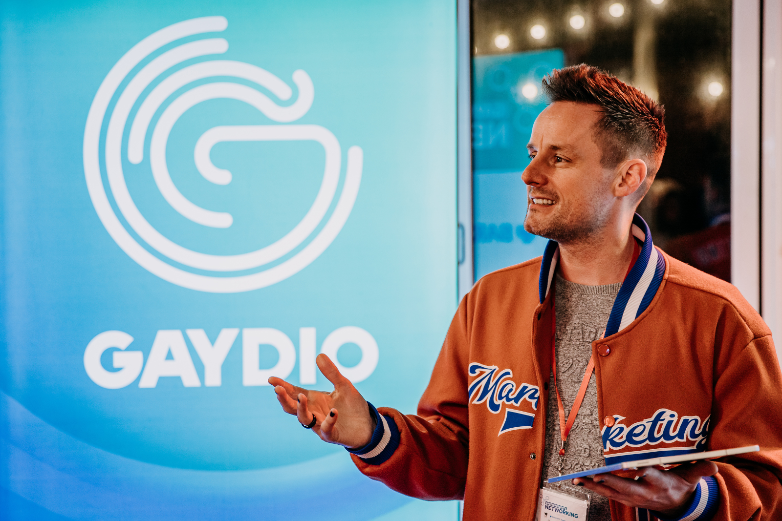 A man smiling and gesturing while standing in front of a large screen displaying the GAYDIO logo, wearing a varsity jacket with the word 'Marketing' on it.