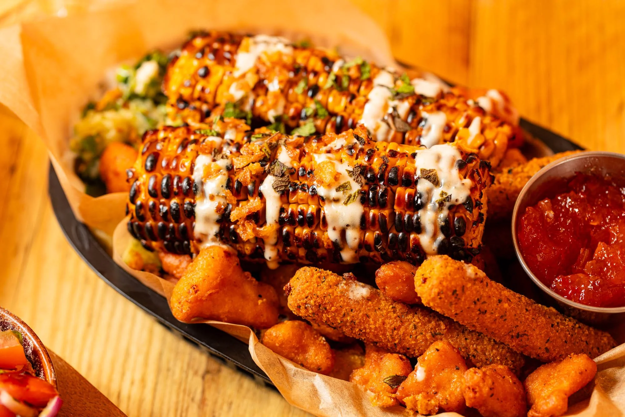 Two grilled corn cobs topped with sauce and herbs, served on a tray with fried chicken, tater tots, and a side of salsa.