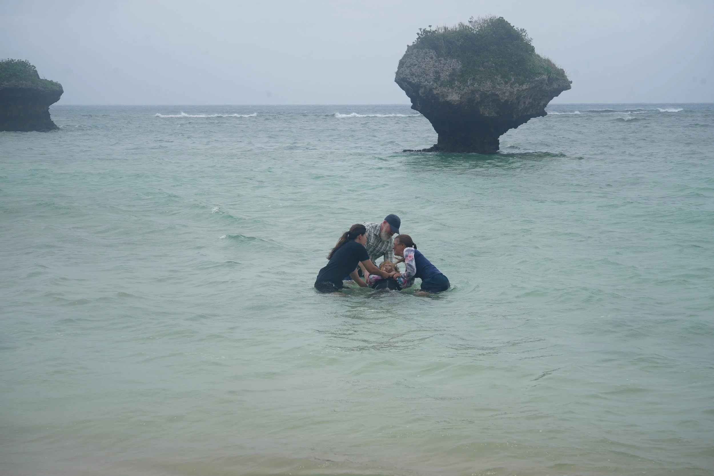 A group of people in the shallow water near the shore, appearing to assist or rescue someone. In the background, there are large sea stacks and a hazy sky.