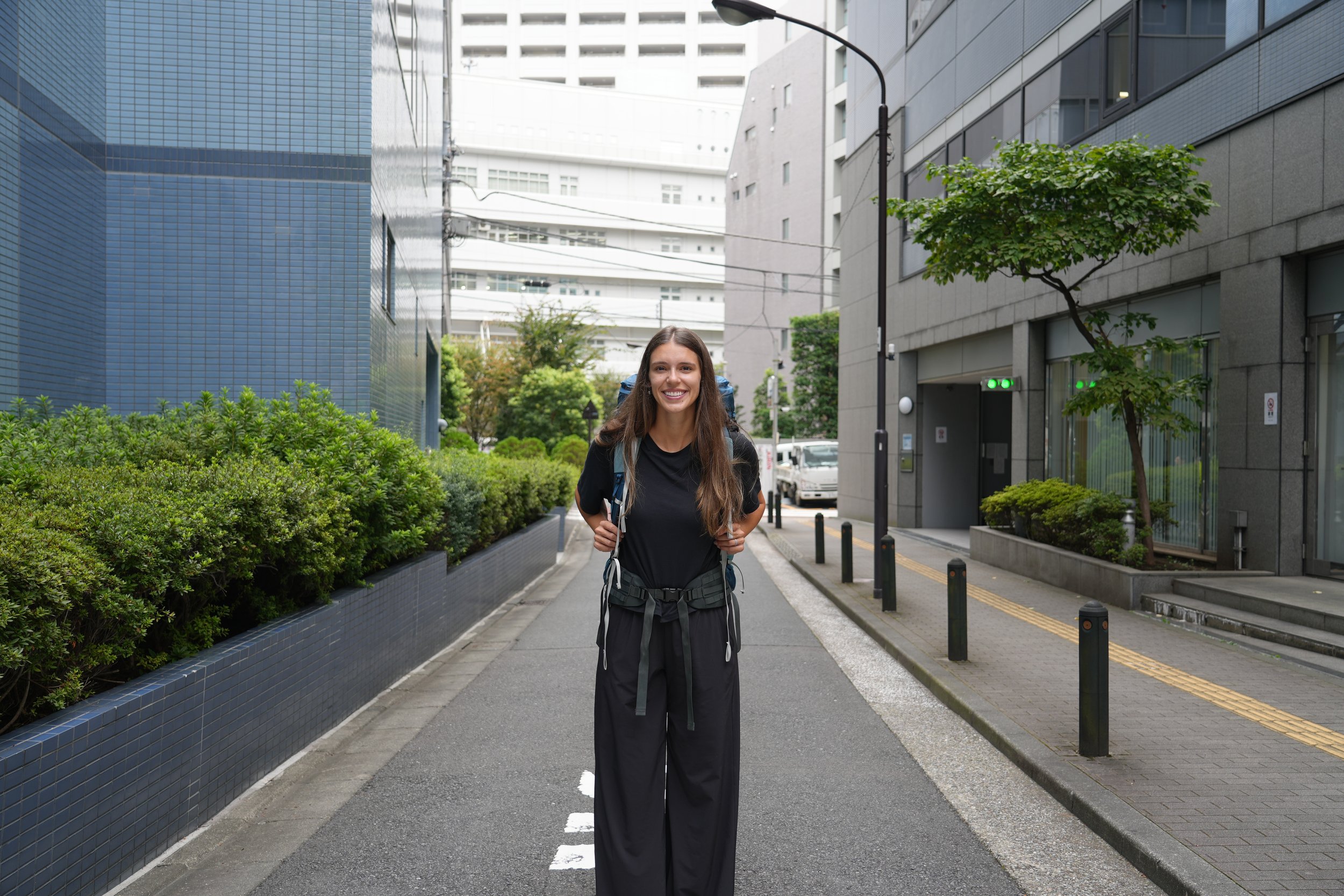 A young woman with long brown hair, wearing a black t-shirt and black pants, smiling with a backpack, walking on a city sidewalk lined with green shrubs and trees, surrounded by modern buildings and urban infrastructure.