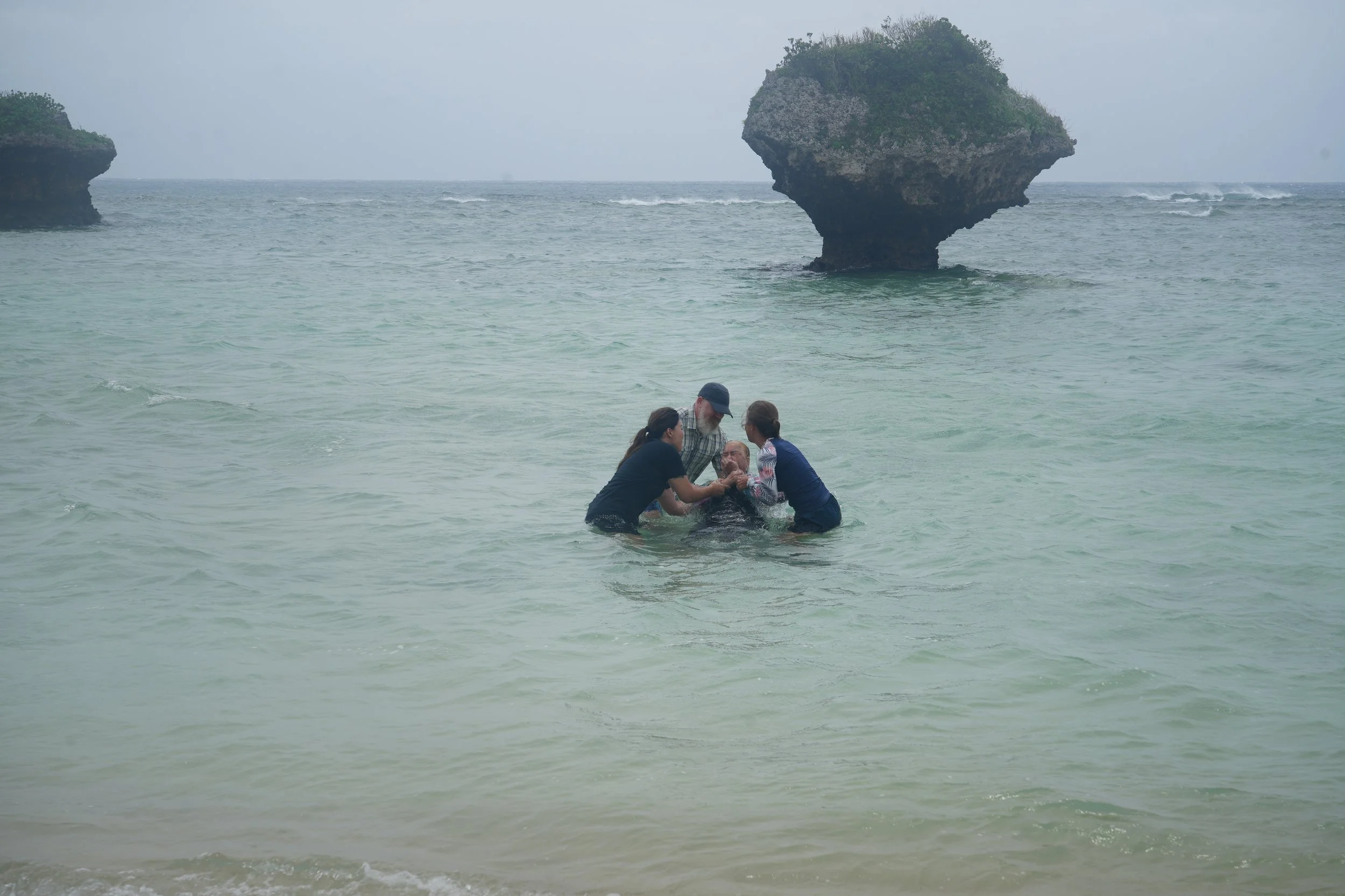 People carry someone in the ocean with large rocky islands in the background.