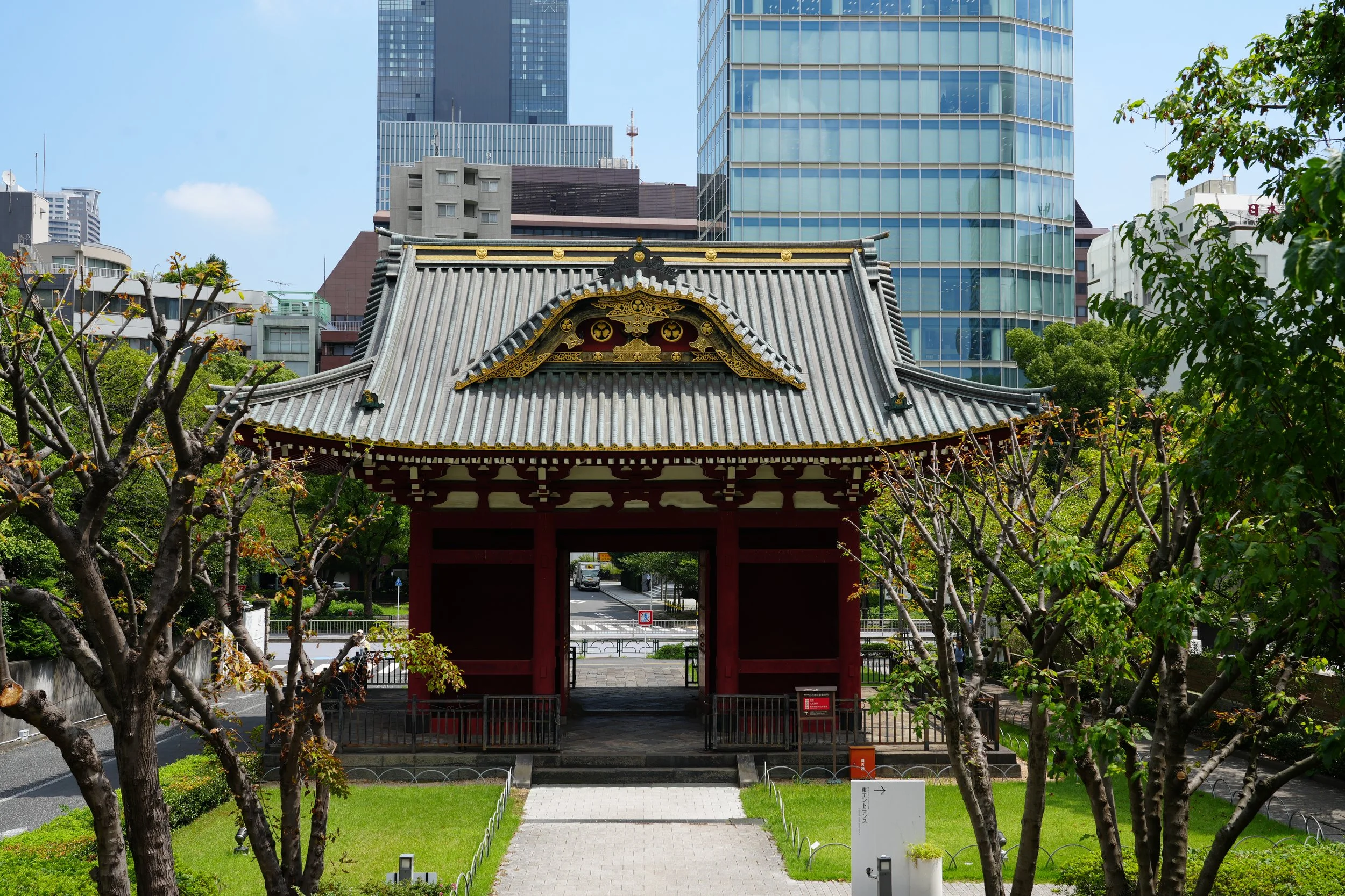 Traditional Japanese shrine gate with curved roof, gold decorative accents, in front of modern tall glass buildings, trees and greenery.