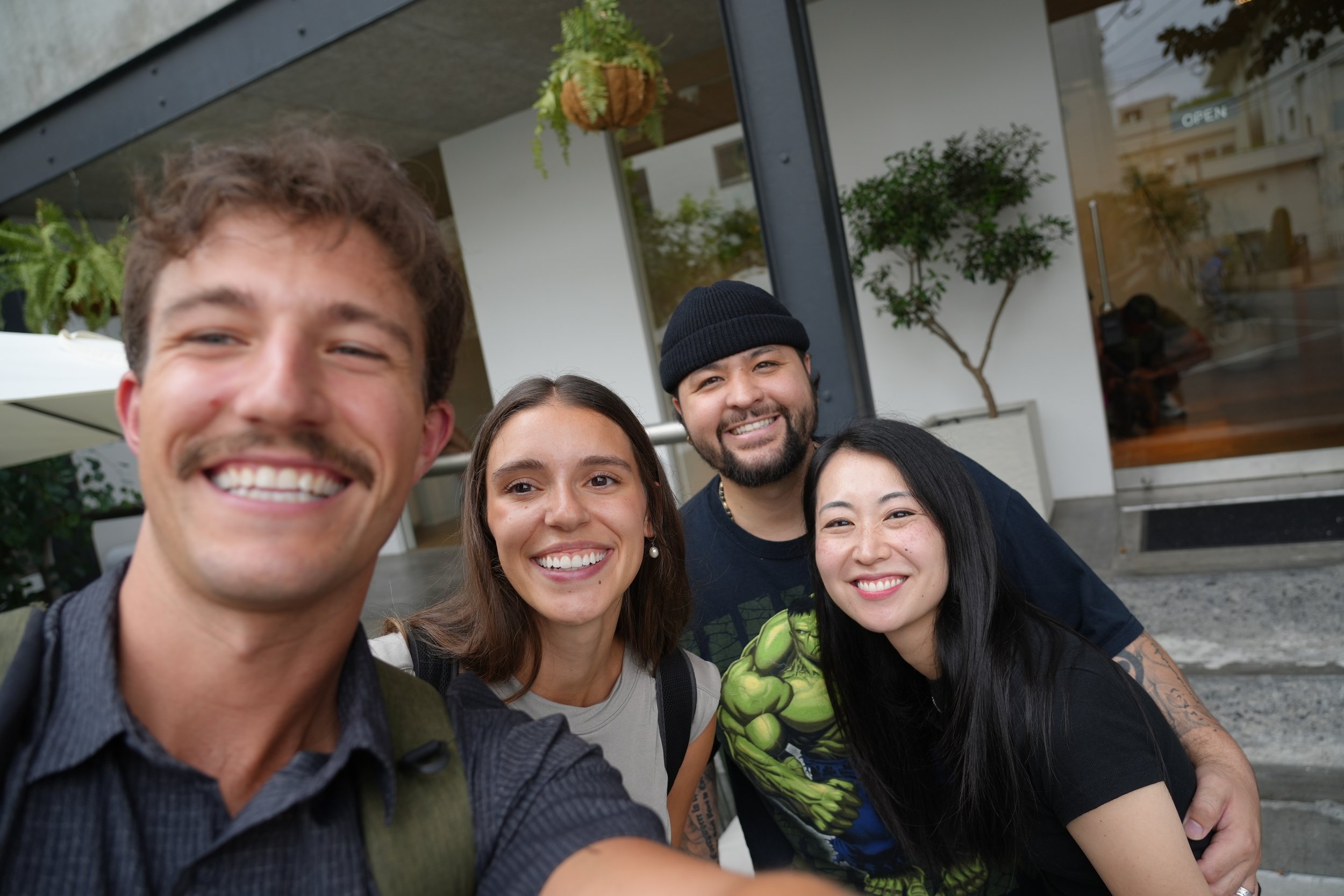 A group of five friends taking a selfie outside of a modern building with plants and large glass windows in the background.