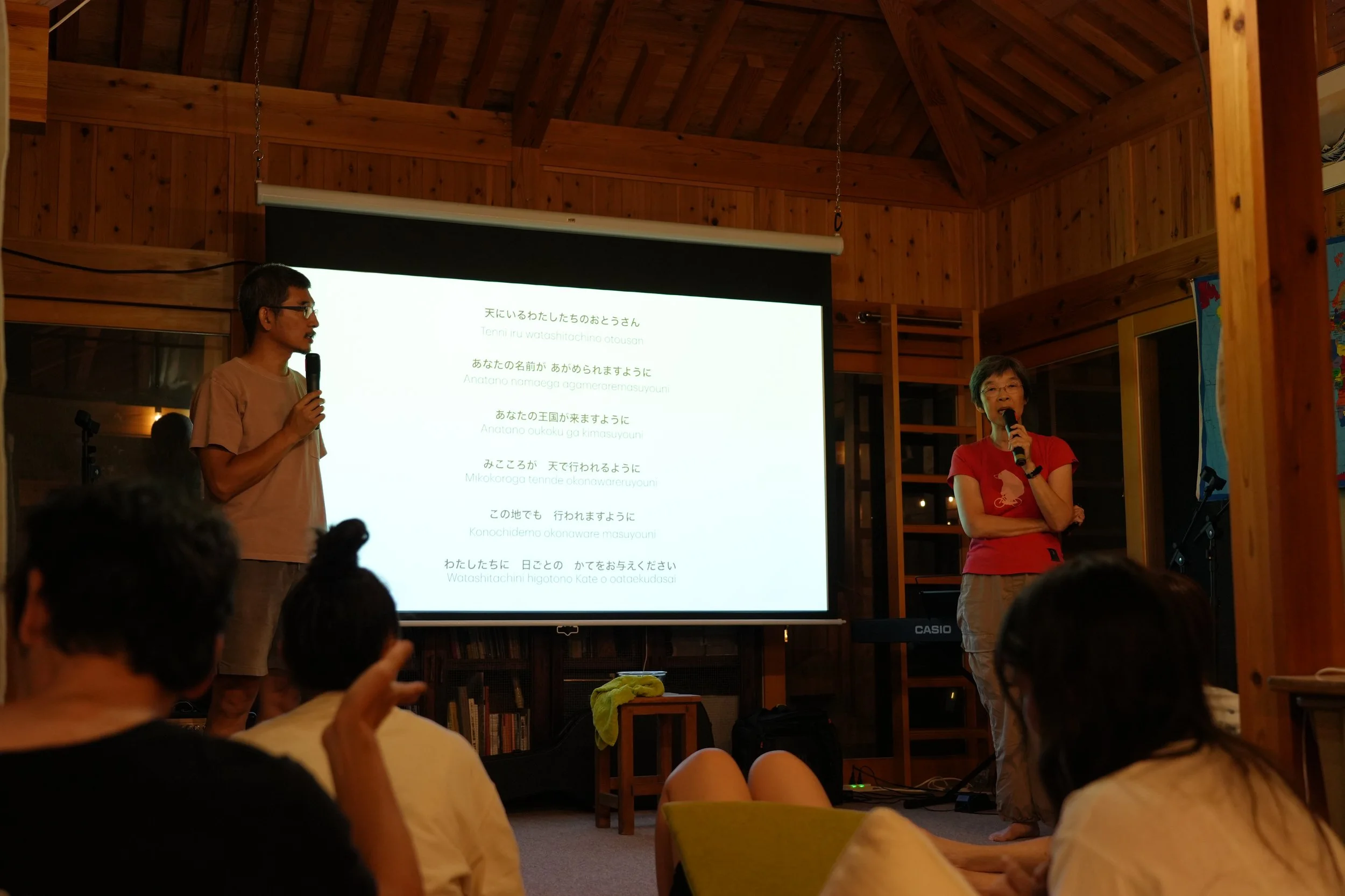 Two people are giving a presentation indoors on a large screen with Japanese text. The man on the left is holding a microphone and wearing glasses and a beige shirt. The woman on the right is holding a microphone and wearing a red shirt. The audience