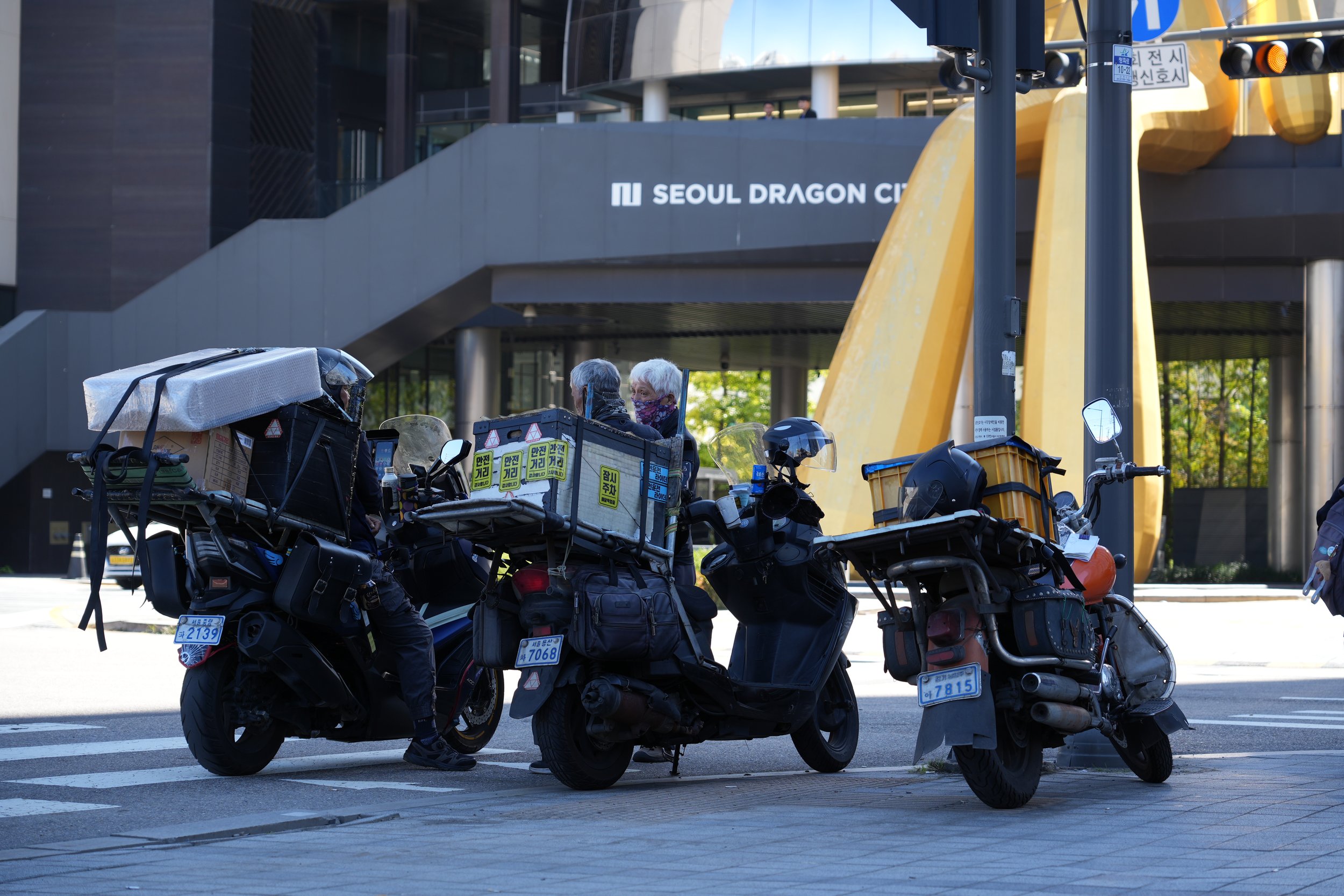 Several delivery motorcycles parked on the street, with two elderly women sitting on one of the motorcycles, and a building with a sign that reads 'Seoul Dragon City' in the background.