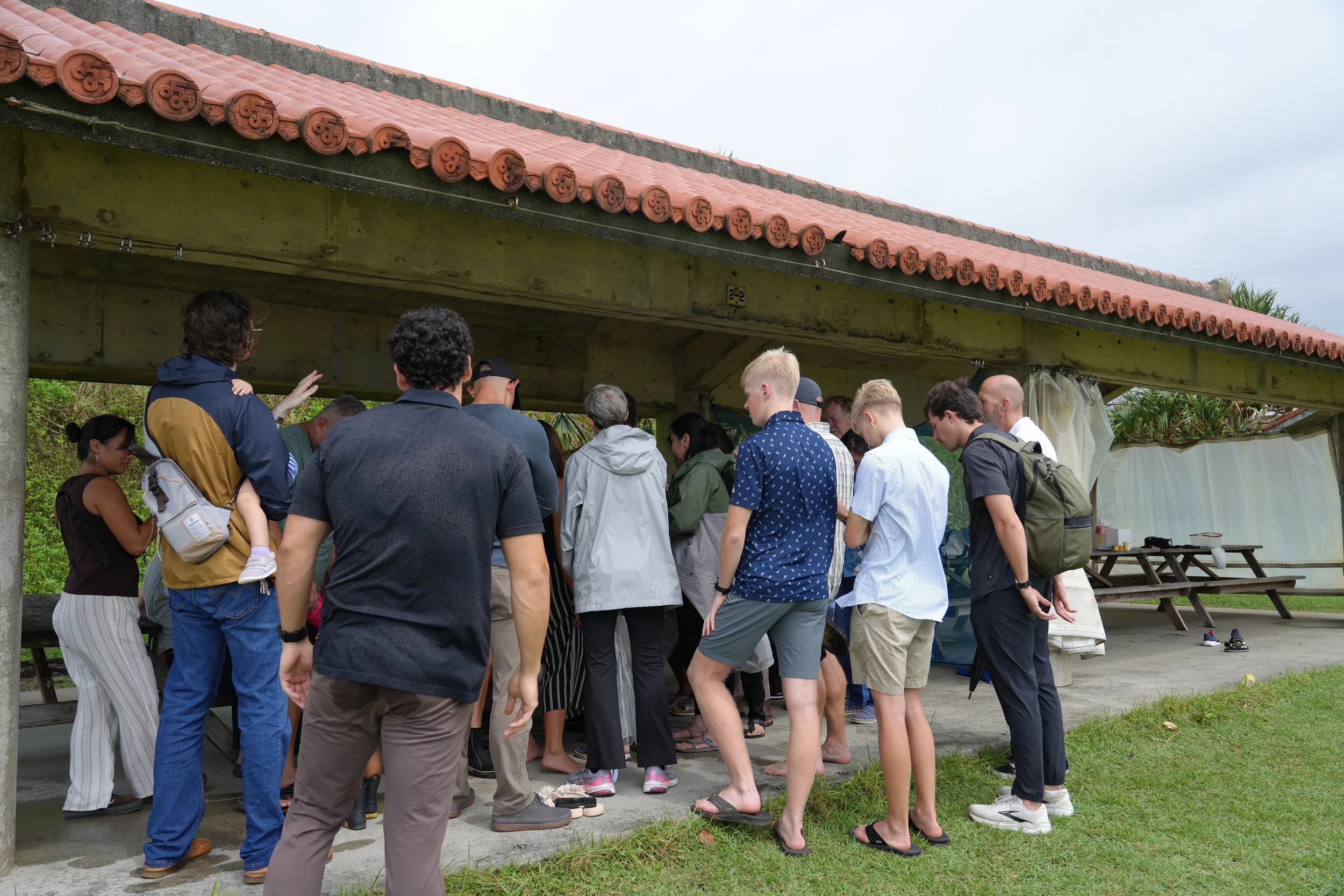 Group of people gathered under a shelter with a traditional tiled roof, outdoors on a cloudy day.