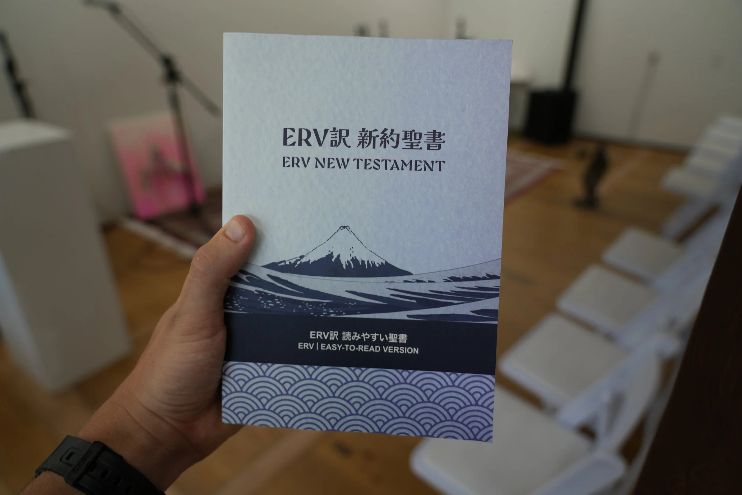 Person holding a booklet titled 'ERV New Testament' with an illustration of Mount Fuji on the cover in a room with white walls and a wooden floor.