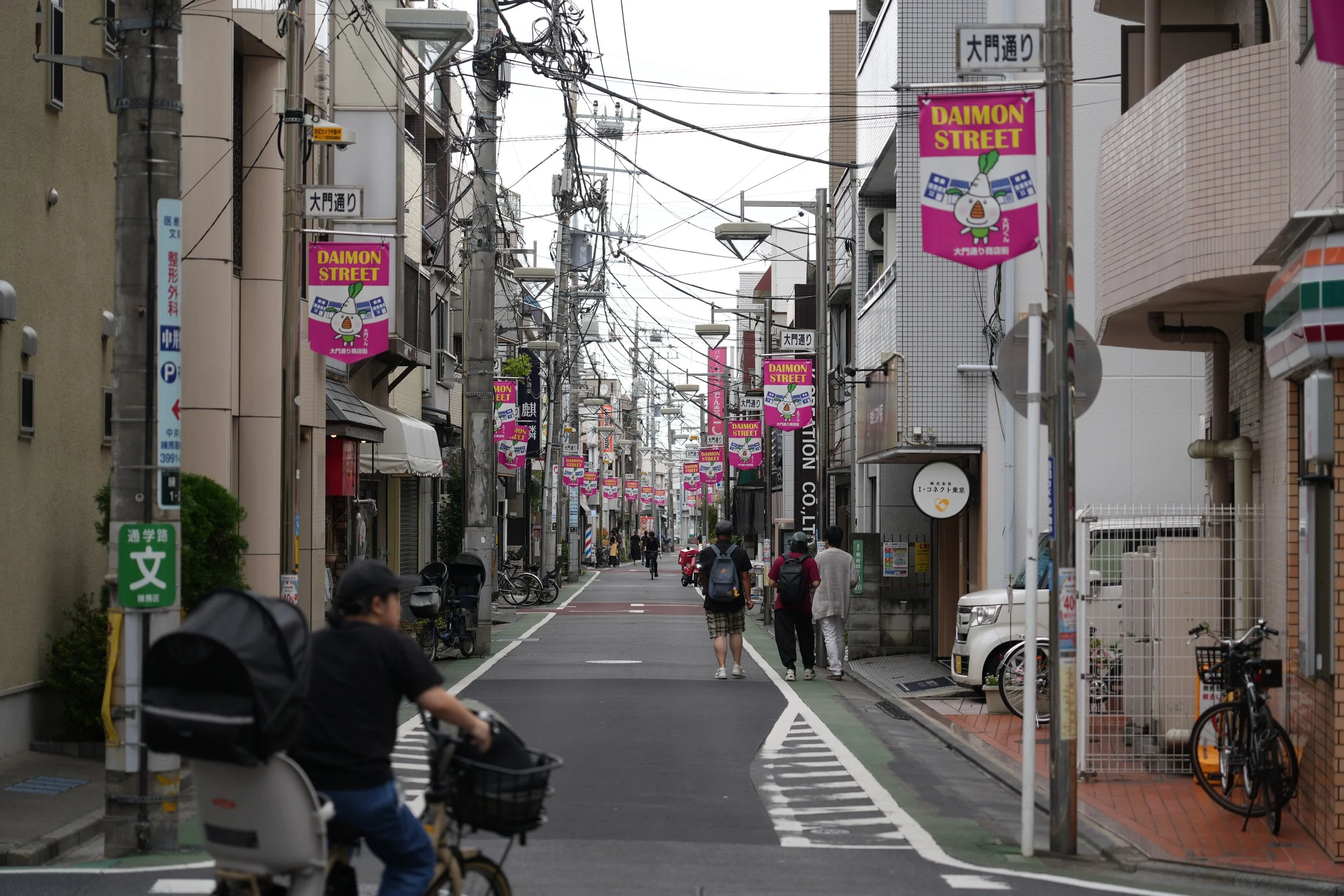 A street scene on Daimon Street in Japan with pedestrians, bicycles, utility poles, and colorful banners hanging above the street.