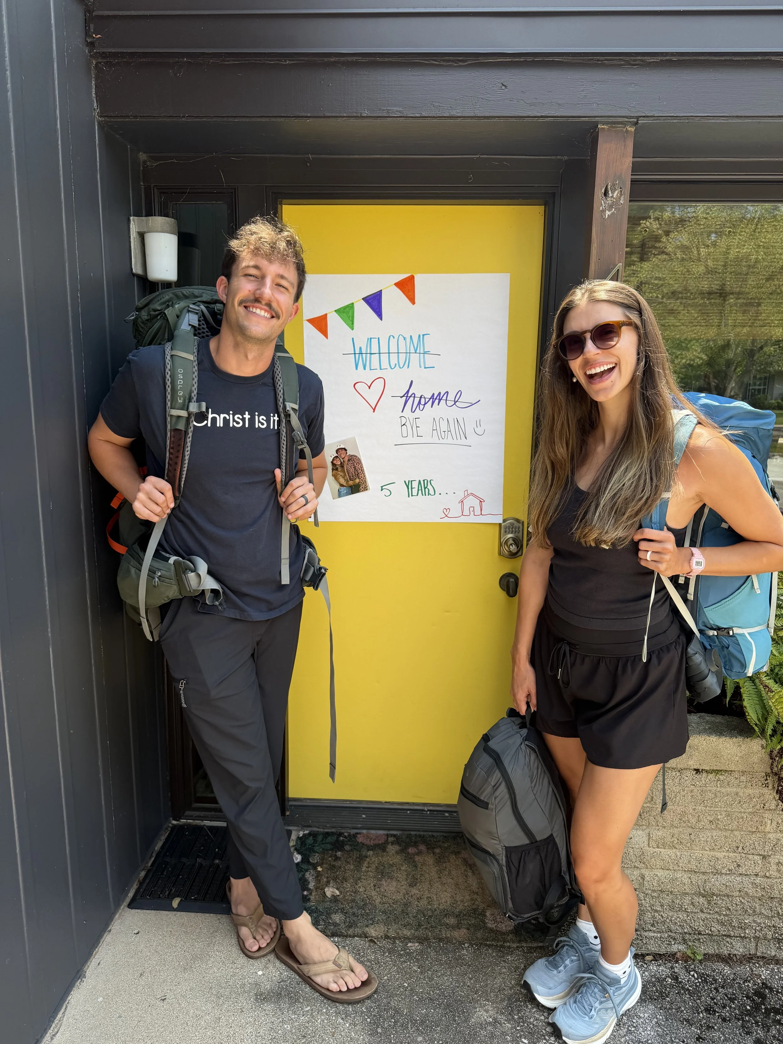 Two smiling travelers, one man and one woman, standing in front of a yellow door with a welcome sign and colorful decorations, carrying backpacks and wearing casual clothing.