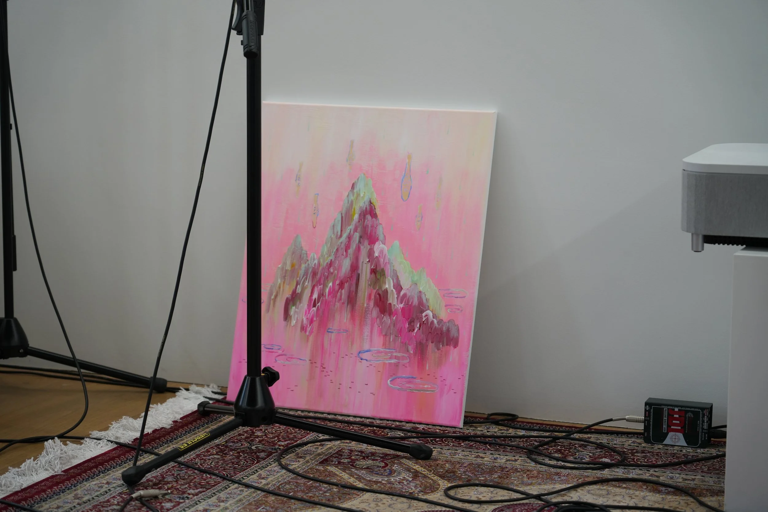 A colorful painting of a mountain scene with pink, green, and purple hues leaning against a plain white wall, with a microphone stand and audio equipment around it on a patterned rug.