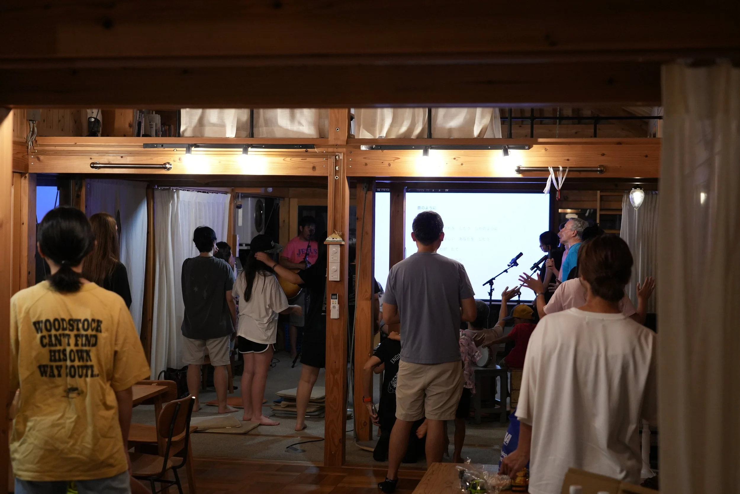 Group of people gathered indoors, some standing and some sitting, watching a presentation on a screen, with wooden beams and curtains in the background.
