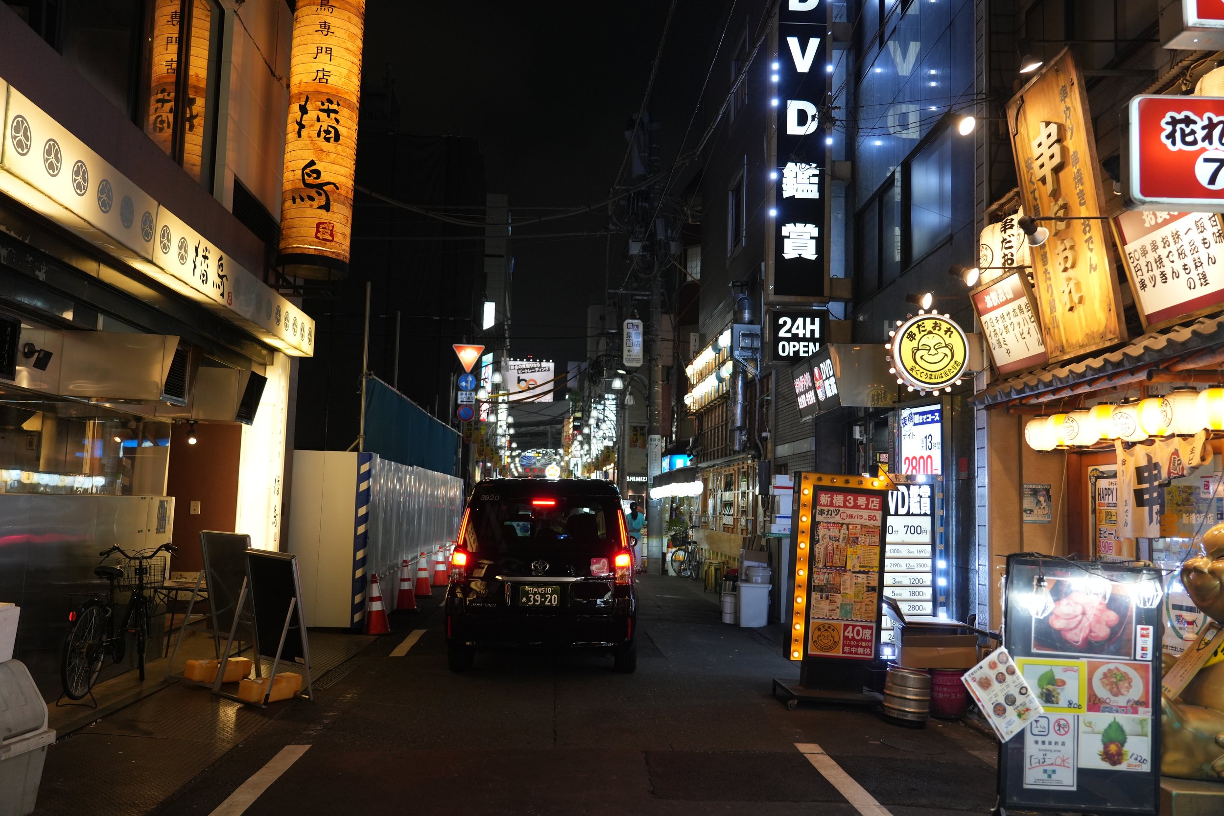 Night street scene in Japan with illuminated signs, paper lanterns, and storefronts on both sides of the street, including a car driving down the narrow road.
