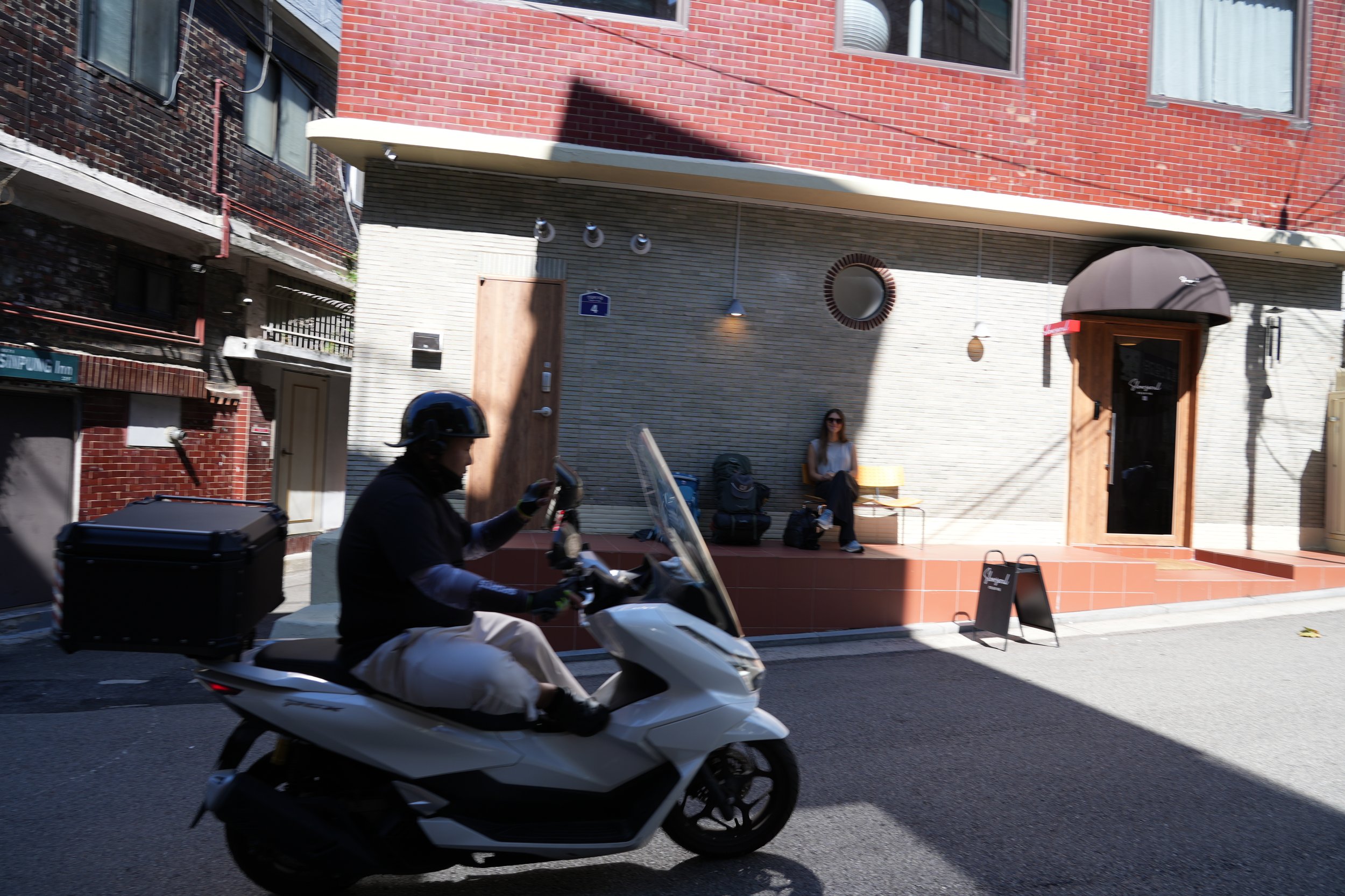 Person riding a white motorcycle with a black storage box on the back, passing in front of a building with a red brick and gray exterior wall, and a woman sitting on a bench outside the building, wearing sunglasses and a sleeveless top.