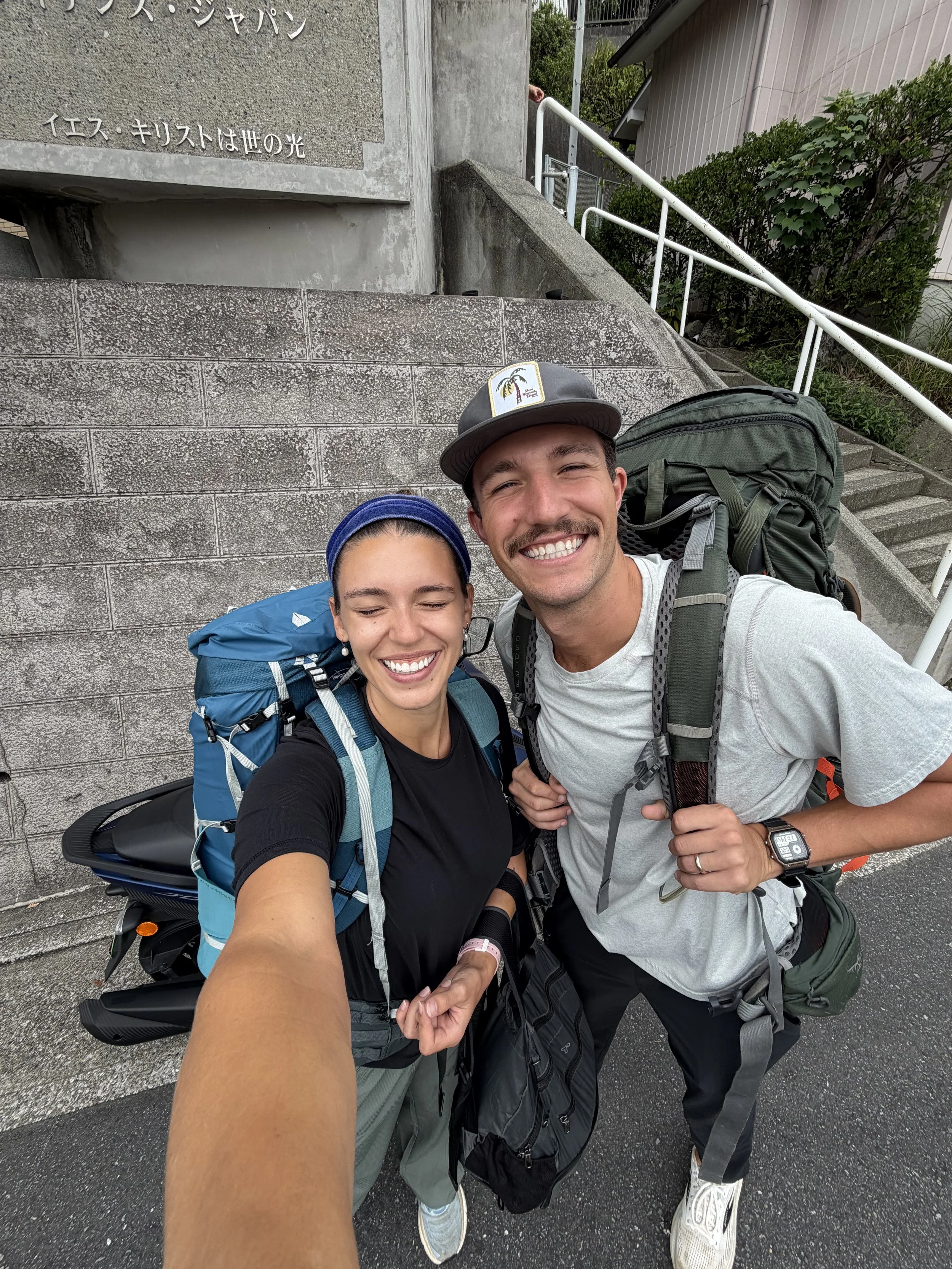 Two hikers taking a selfie in front of a concrete building with stairs and greenery. Both are smiling, wearing backpacks and outdoor clothing.