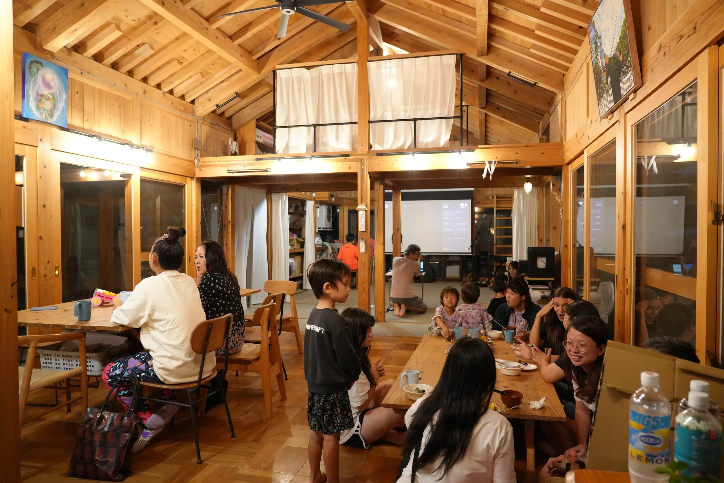 Indoor gathering of people in a wooden-finished room with a high vaulted ceiling, some standing and some seated at tables, children and adults chatting, with paintings on the walls and a projector screen in the background.