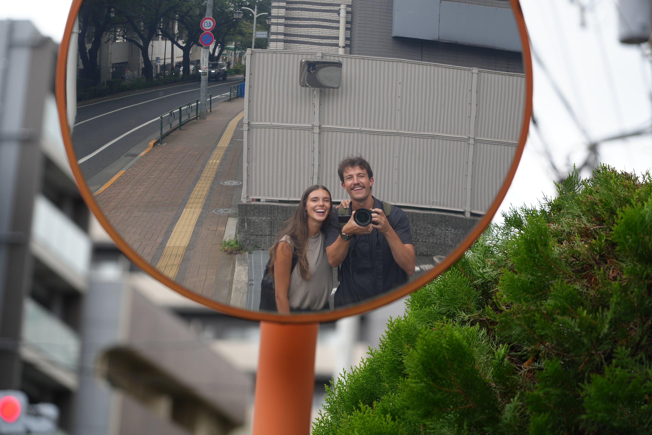 Two people taking a selfie in a convex mirror on a city sidewalk, with parked cars, trees, and buildings in the background.