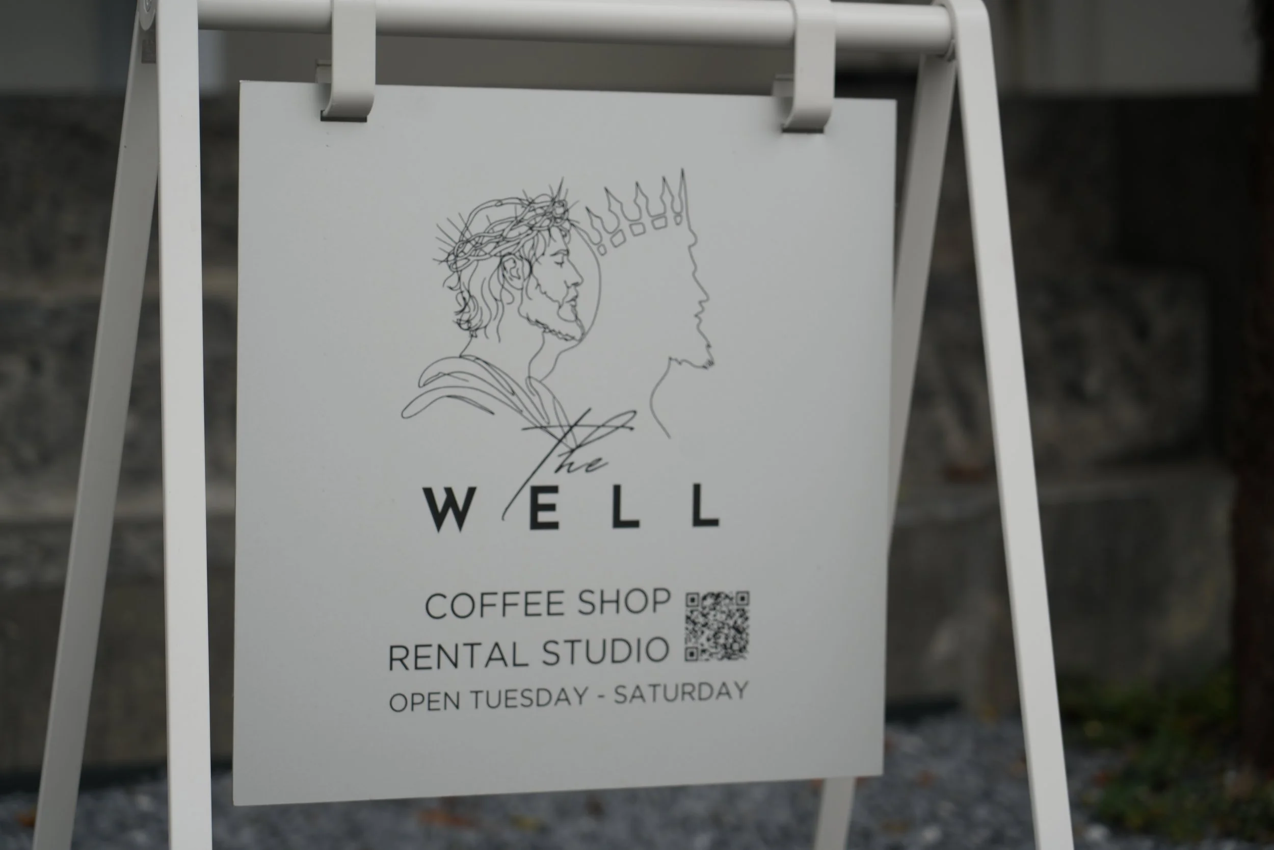 A white A-frame signboard with a line drawing of Jesus Christ wearing a crown of thorns, and the profile of a person's face with a crown of exalted symbols, with the text 'WELL COFFEE SHOP' and details about rental studio and opening days.