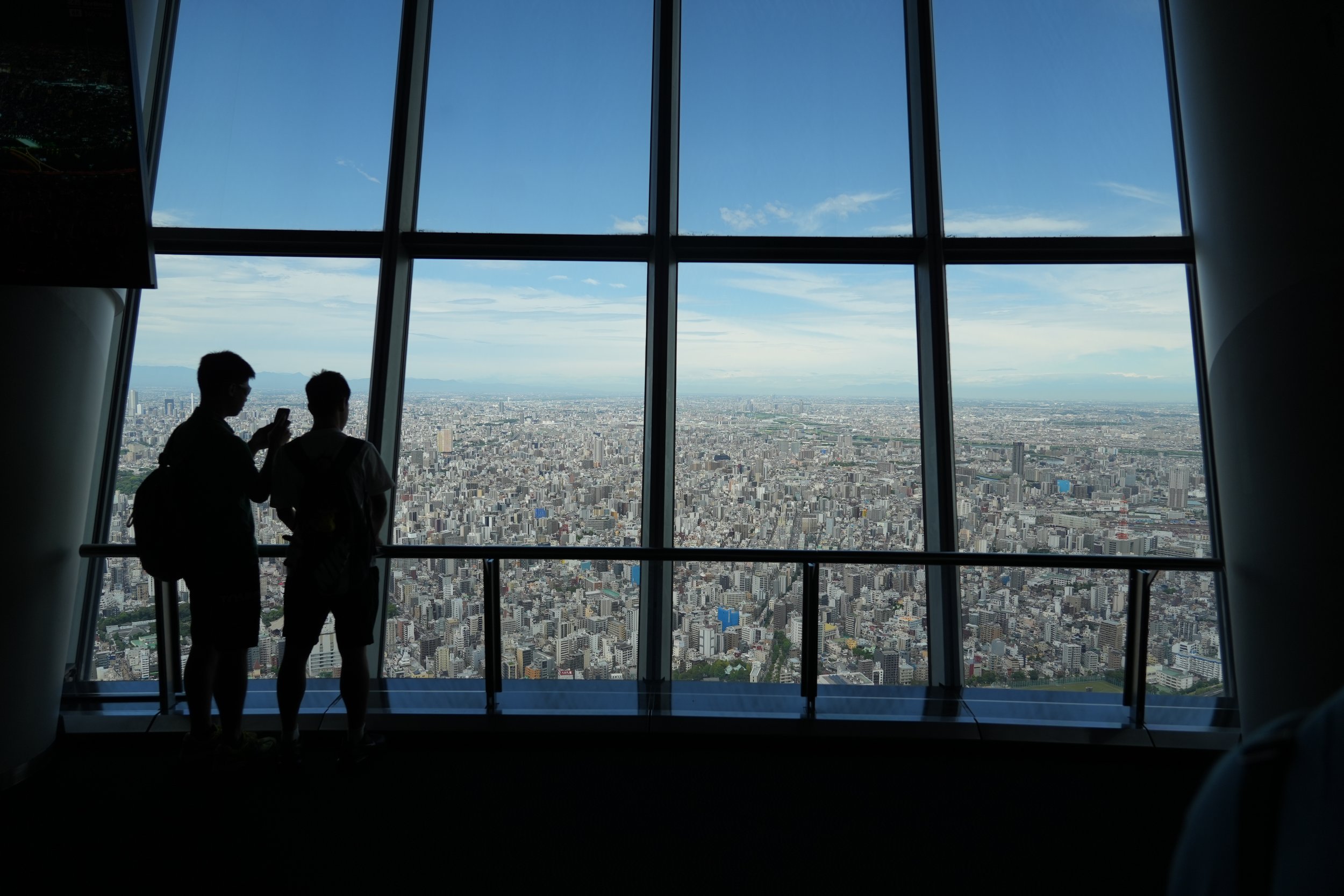 Two people standing inside a tall building, looking out of large windows at a city skyline with many buildings and a clear sky.