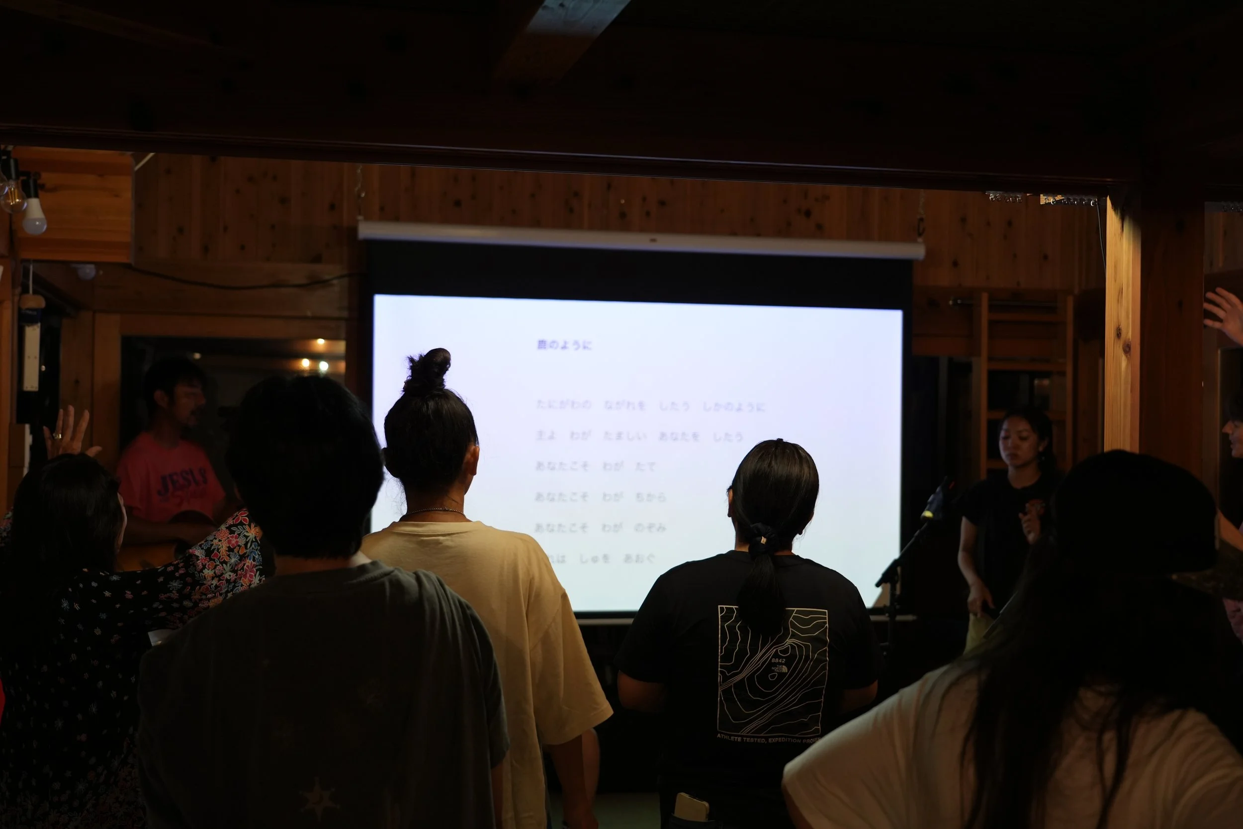 People attending a presentation or lecture in a wooden-furnished room, facing a large screen displaying text.
