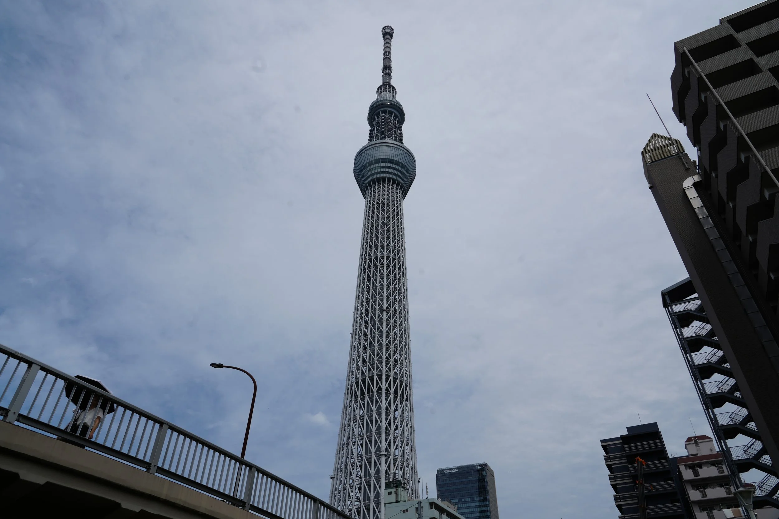 A tall tower with a lattice structure, likely a telecommunications or observation tower, with a rounded observation area near the top and multiple antennas on its pinnacle. It's viewed from a surrounding urban area with modern buildings and a curved 