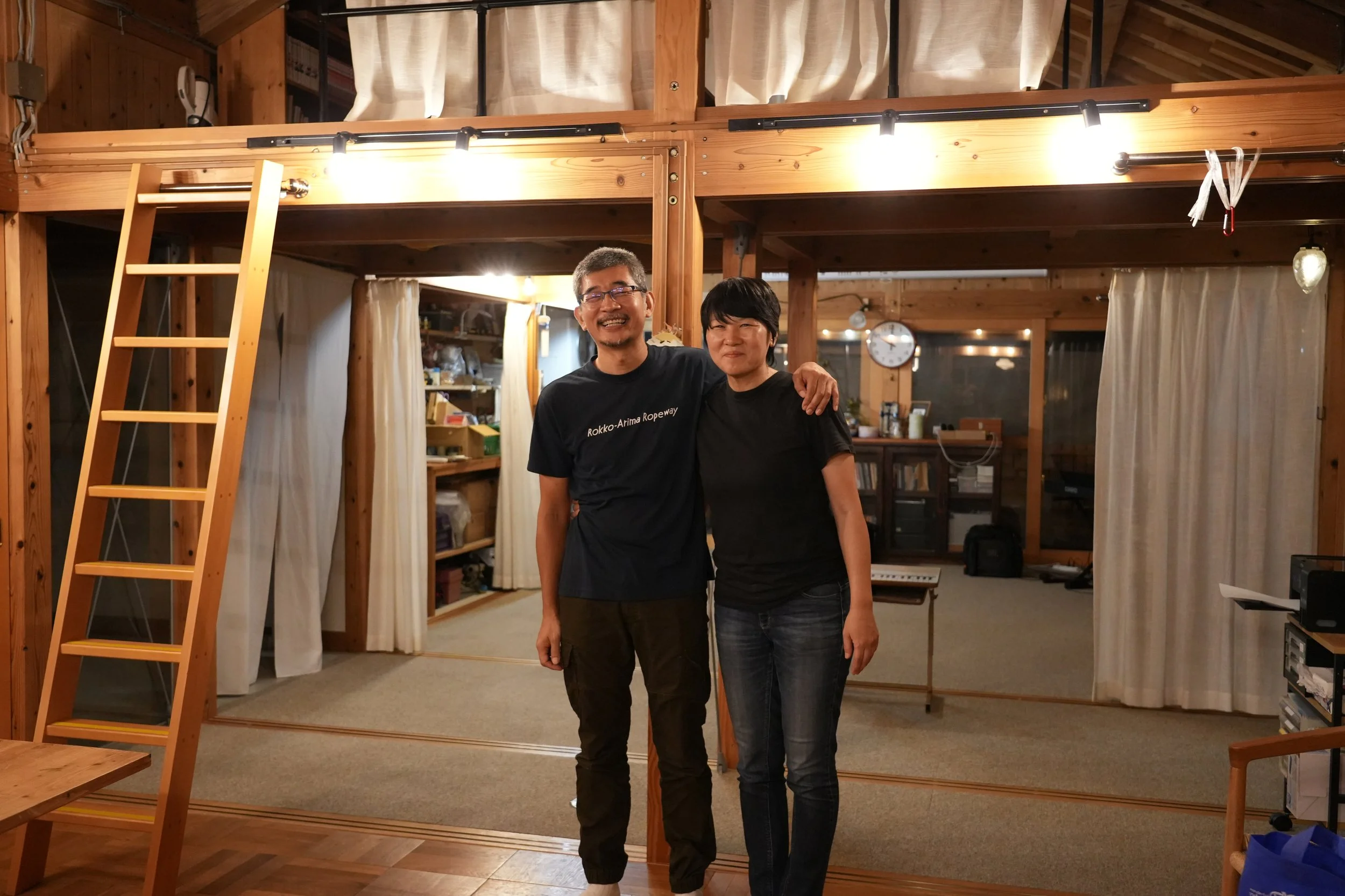 Two smiling men standing inside a wooden room with large mirror and loft area, one with arm around the other, in a cozy, wood-paneled space.