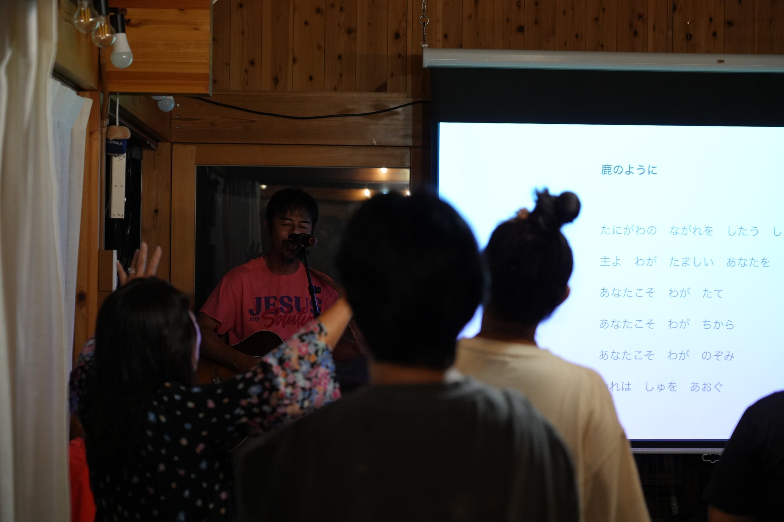 A man singing and playing guitar in front of a projected screen with Japanese text, inside a wooden-paneled room with audience members.