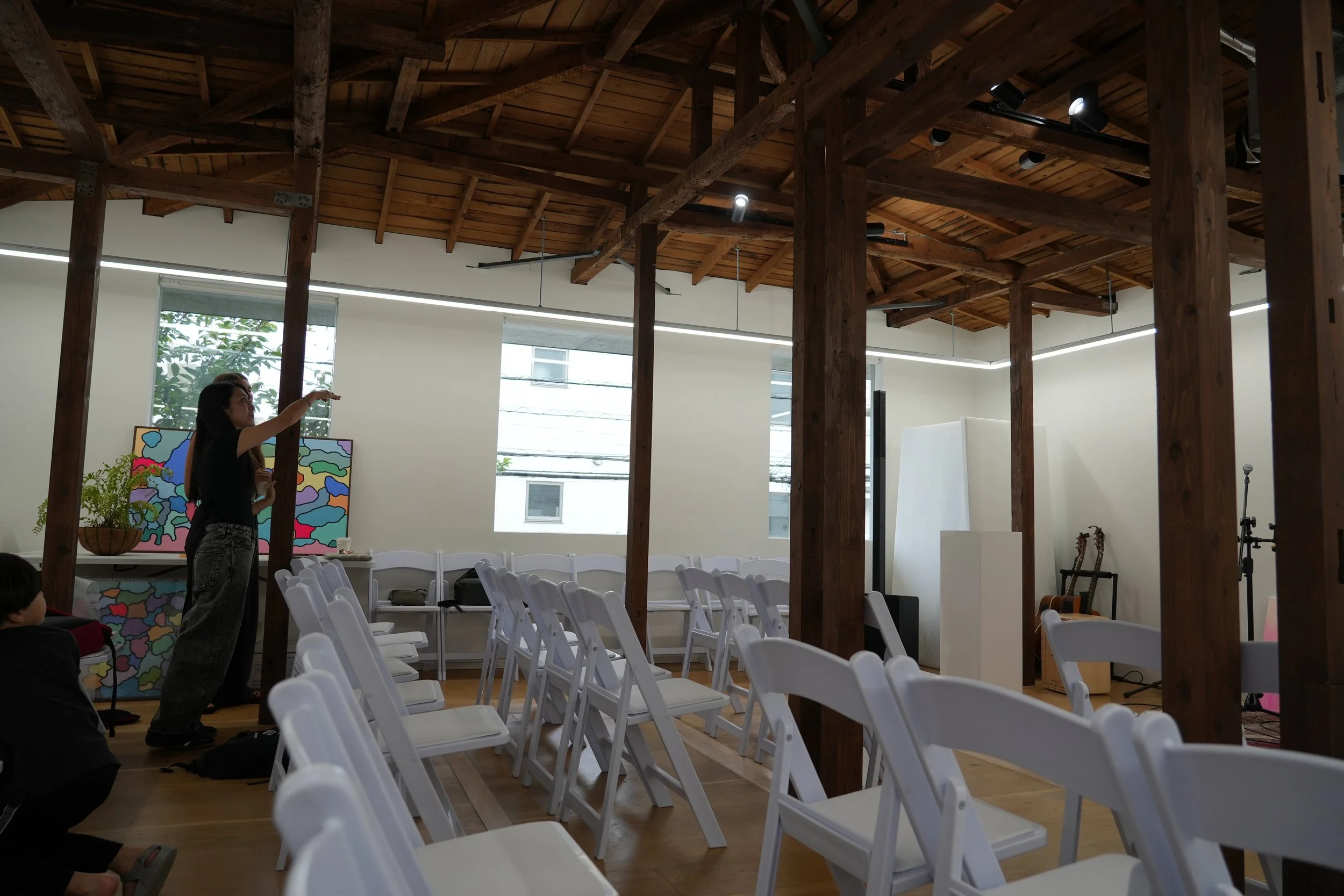 An indoor event space with wooden beams and a high ceiling. Rows of white chairs are arranged, and a woman is standing near a colorful abstract painting, gesturing with her arm. Large windows let in natural light.